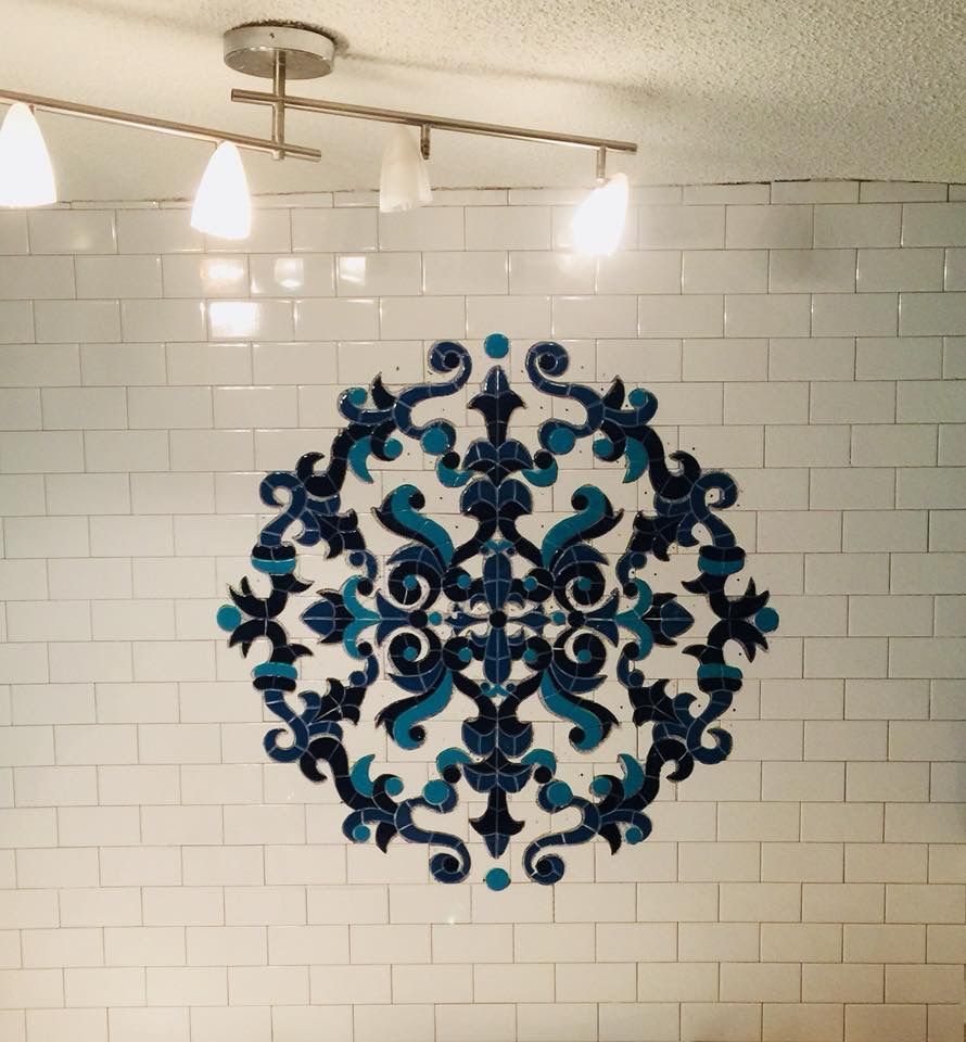 White tiled wall with a dark blue and teal ornate tile medallion. Overhead lighting.