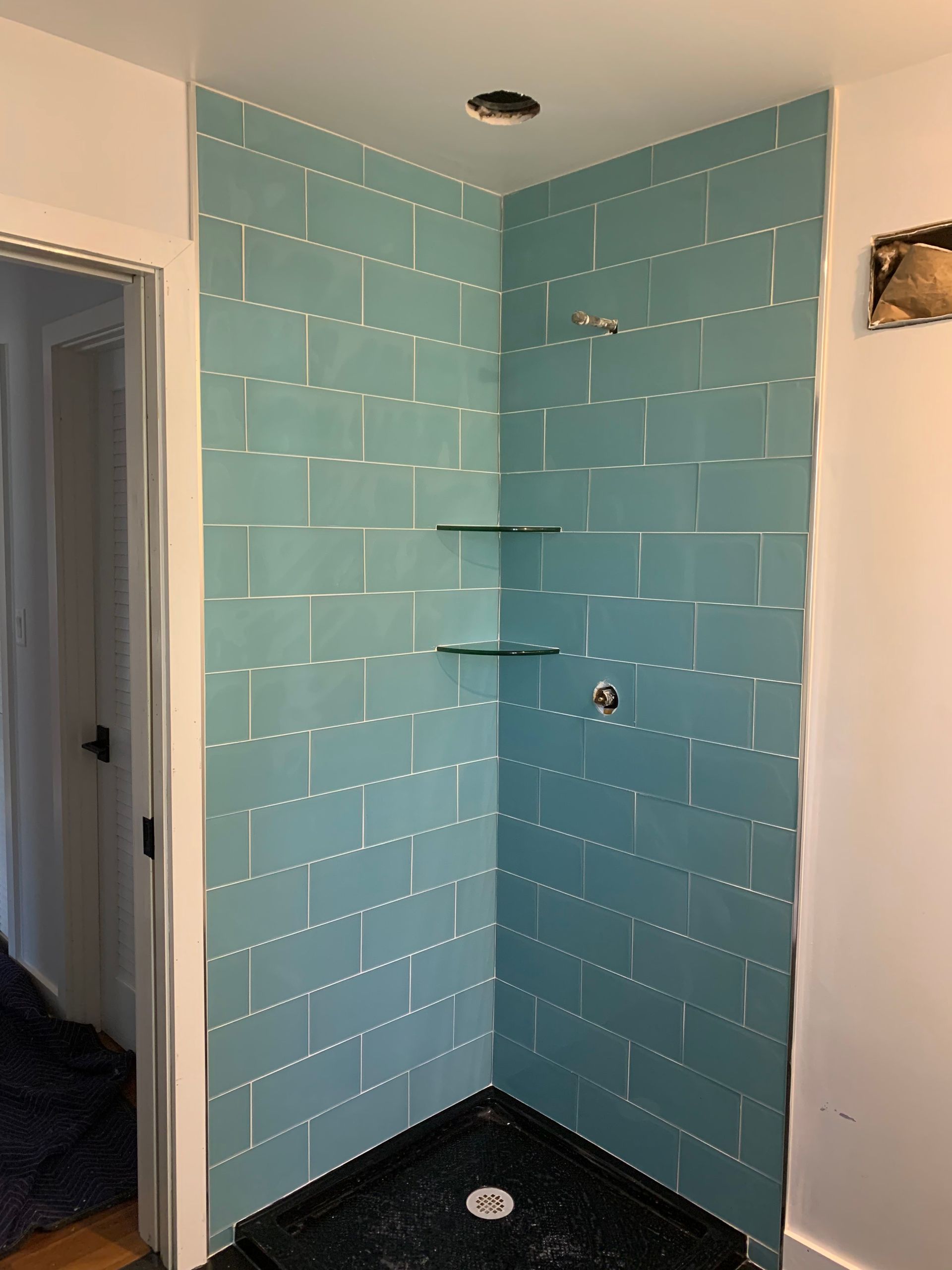 Blue tiled shower corner with glass shelves, black base, and a recessed ceiling light.