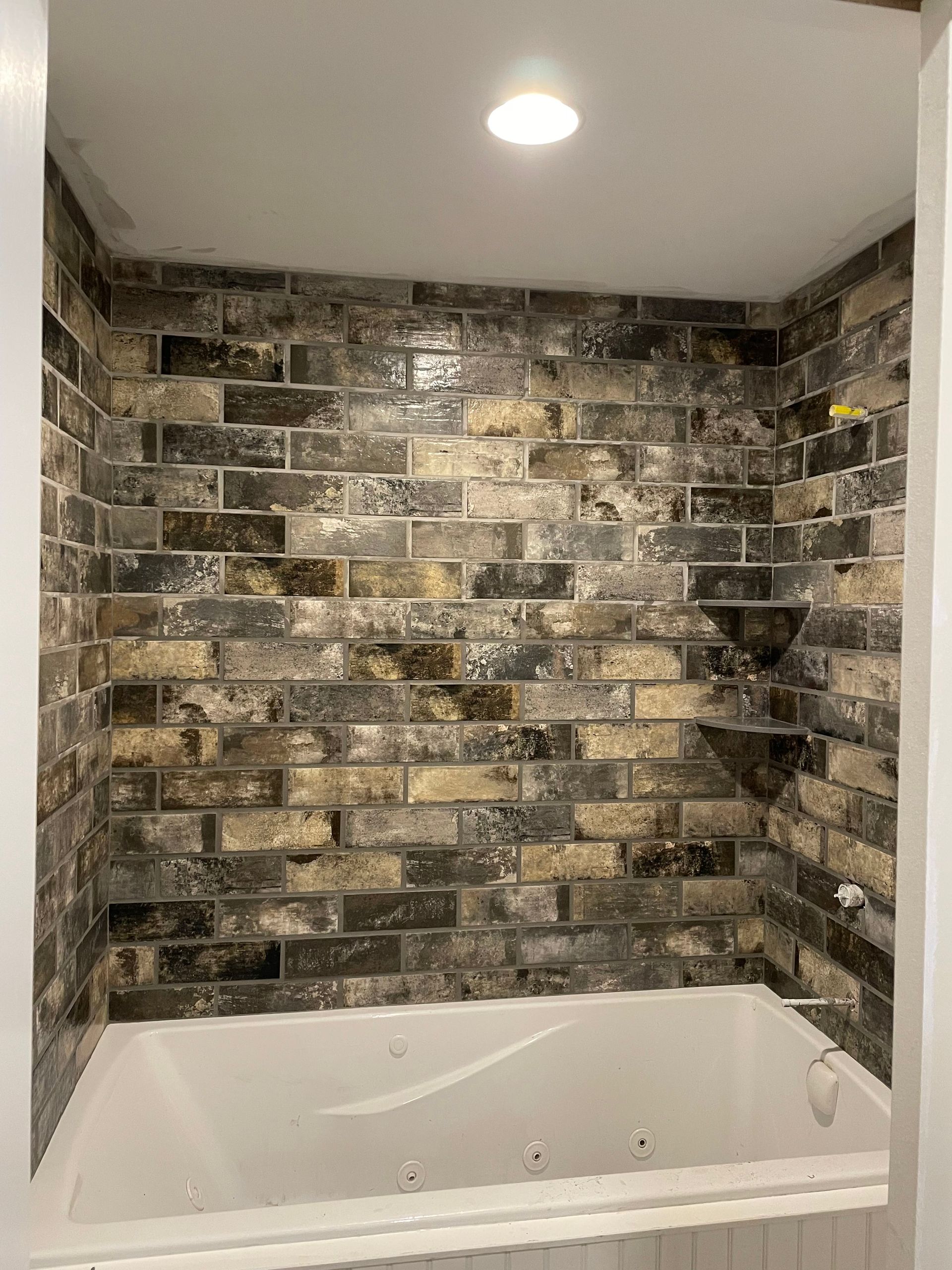 Bathroom with tub and brick-like tile in shades of gray, brown, and gold on the walls. White trim and ceiling.
