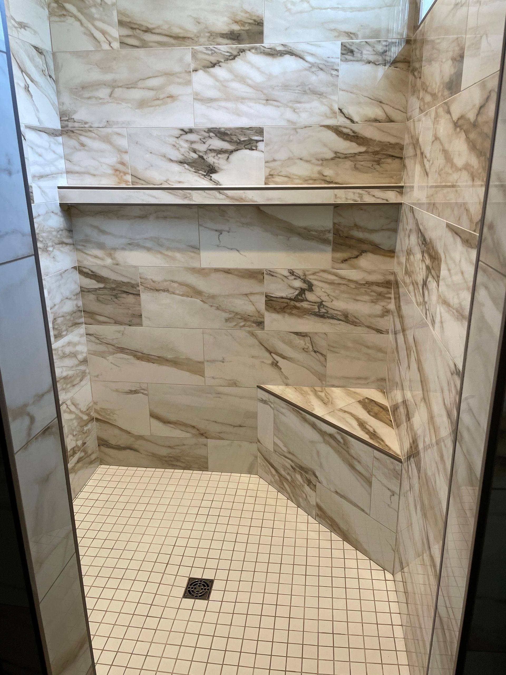 Tiled shower with marble-look walls, a shelf, and a small bench. White mosaic floor with drain.