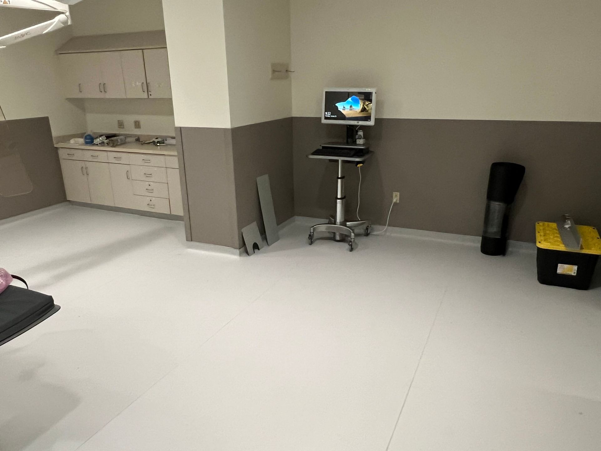 Empty medical room with white floor, gray walls, cabinets, monitor, equipment, and a toolbox.