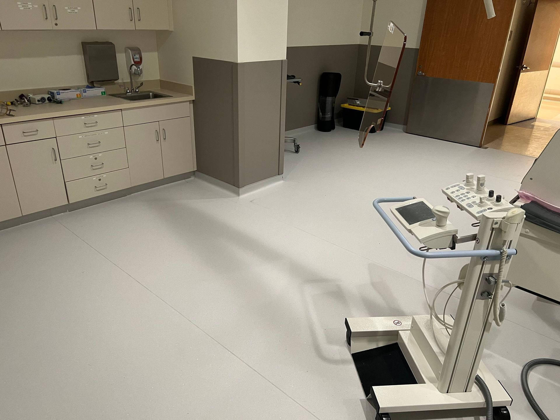 Empty medical room with white flooring, cabinets, and a medical device on wheels.
