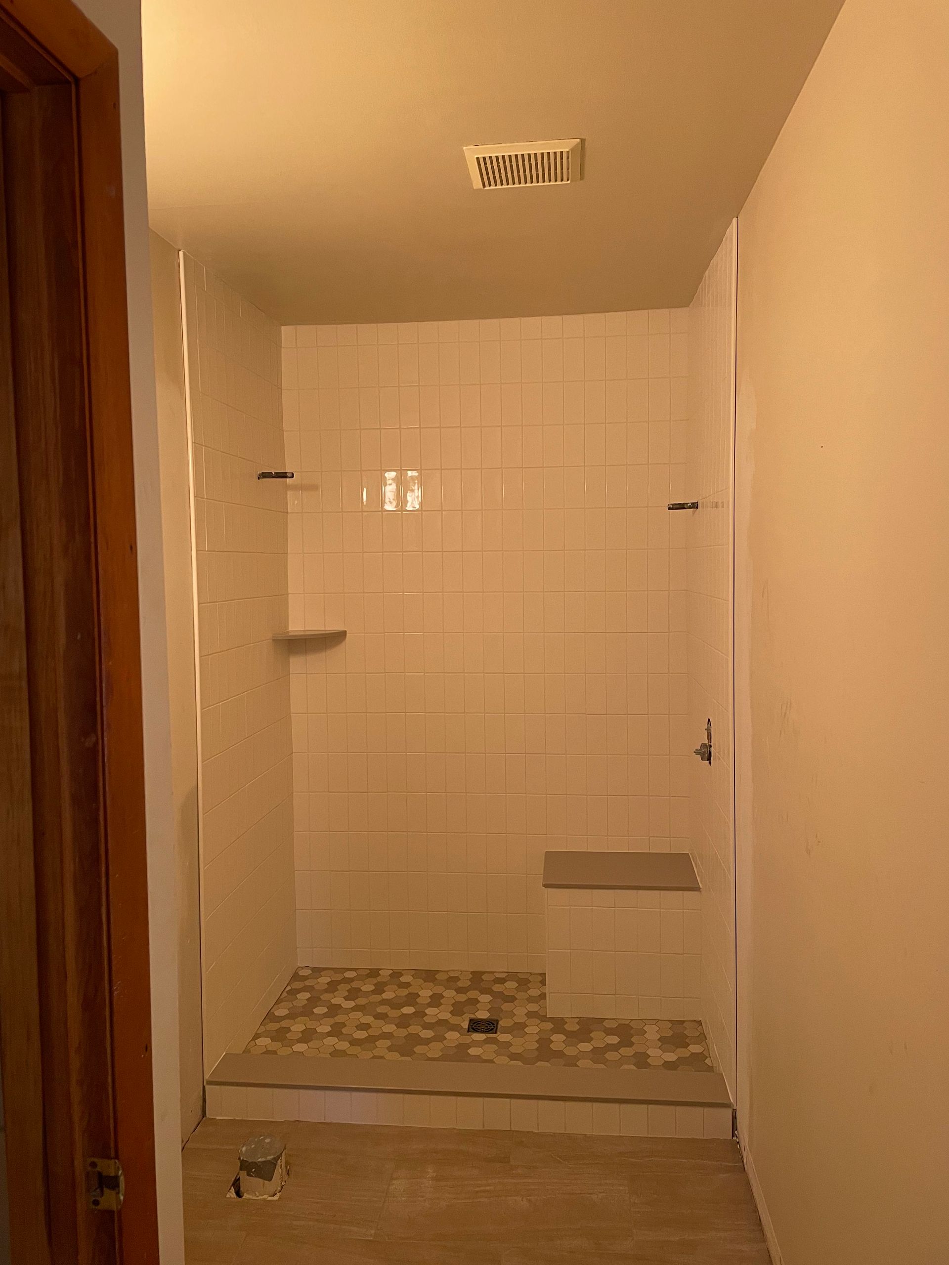 Bathroom under construction. White tiled shower with stone floor, small bench, and recessed shelf.