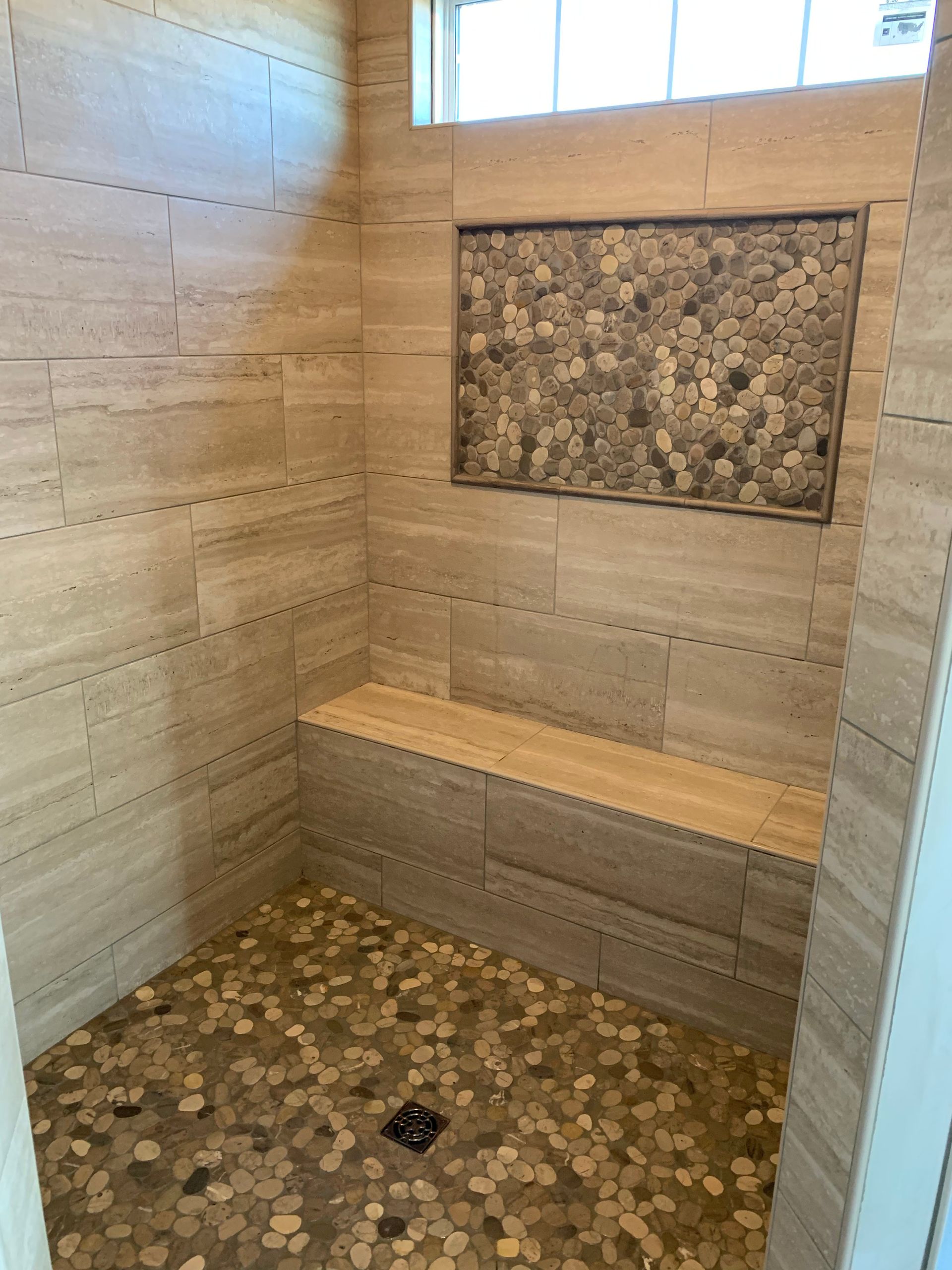Shower with stone tile walls, pebble floor, and wooden bench. Pebble accent in wall niche.