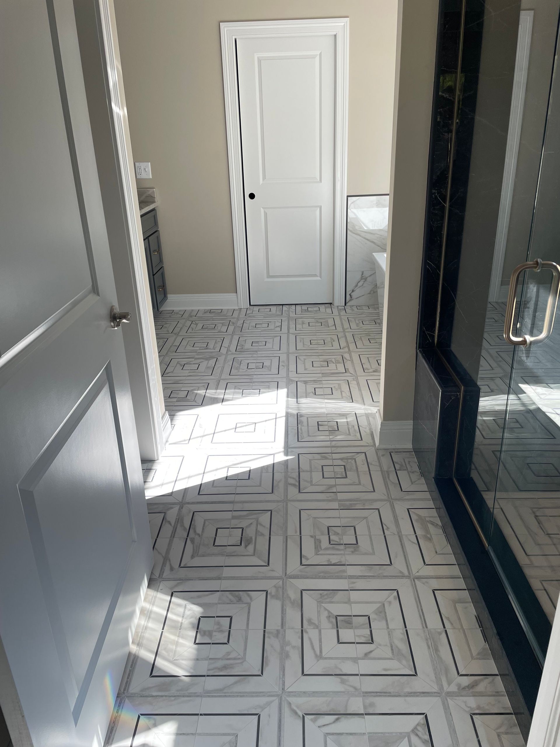Bathroom hallway with white and gray patterned tile floor, white doors, and sunlight.