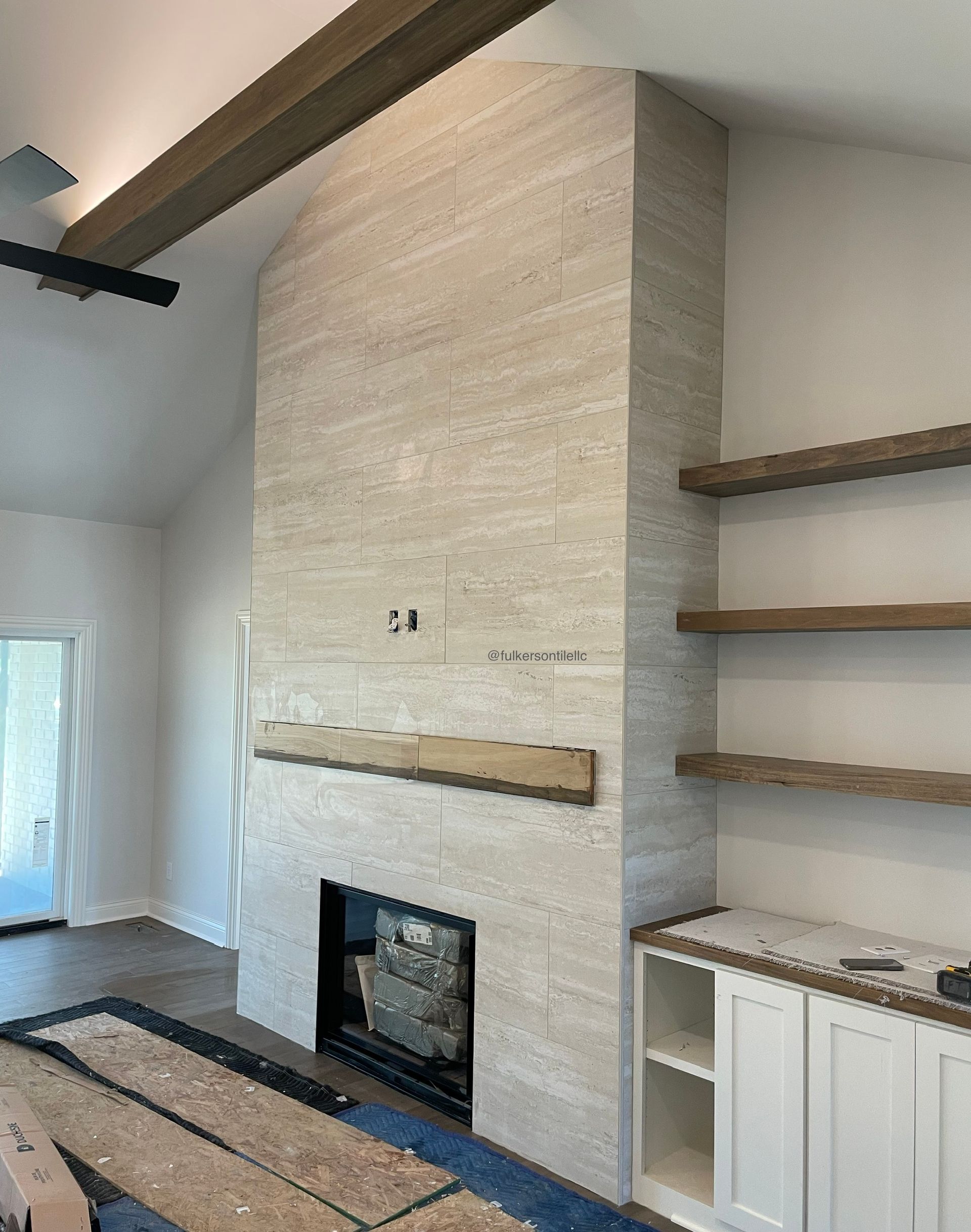 Fireplace with light stone facade, wooden beam mantel and floating shelves, in a room with white walls.