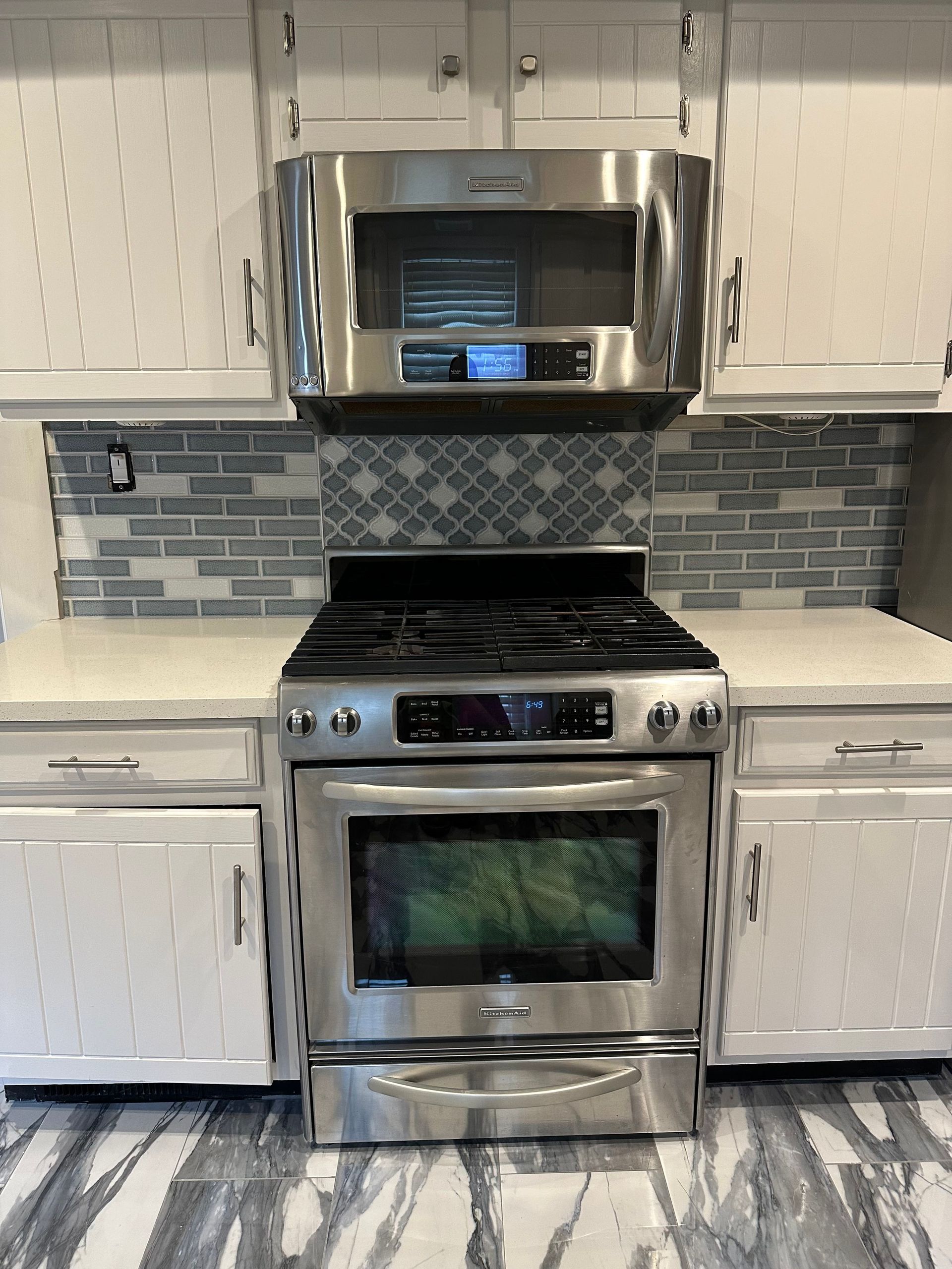 Stainless steel oven and microwave in a kitchen with white cabinets, gray backsplash, and gray and white floor.
