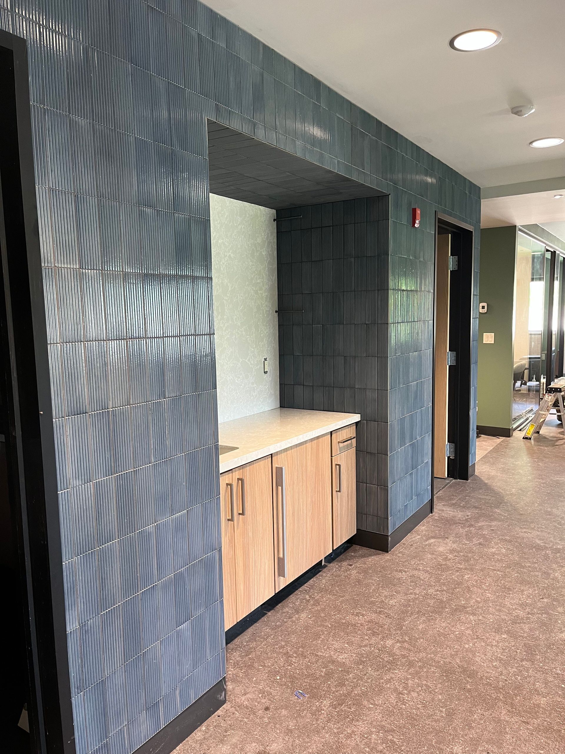 Blue tiled alcove with wood cabinetry and a beige countertop. A hallway is visible to the right.