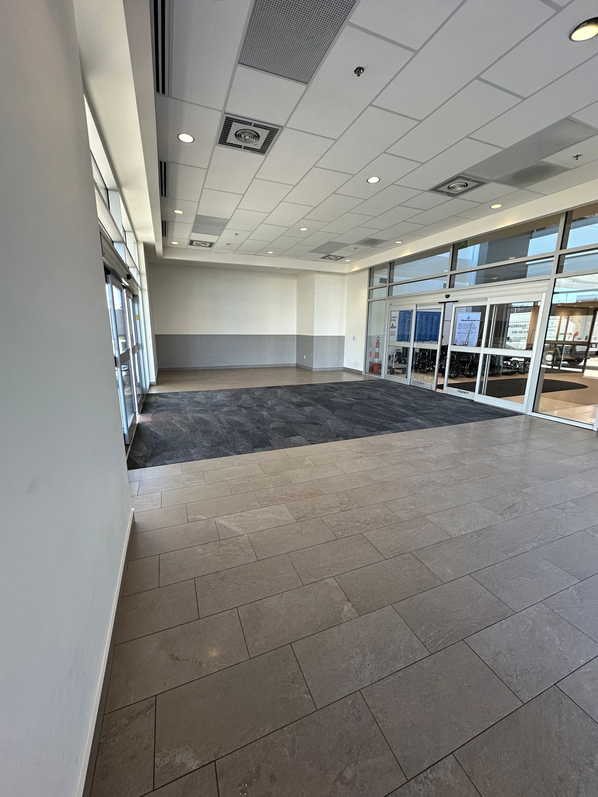 Empty store interior with tile flooring, dark gray carpet, glass doors, and recessed lighting.