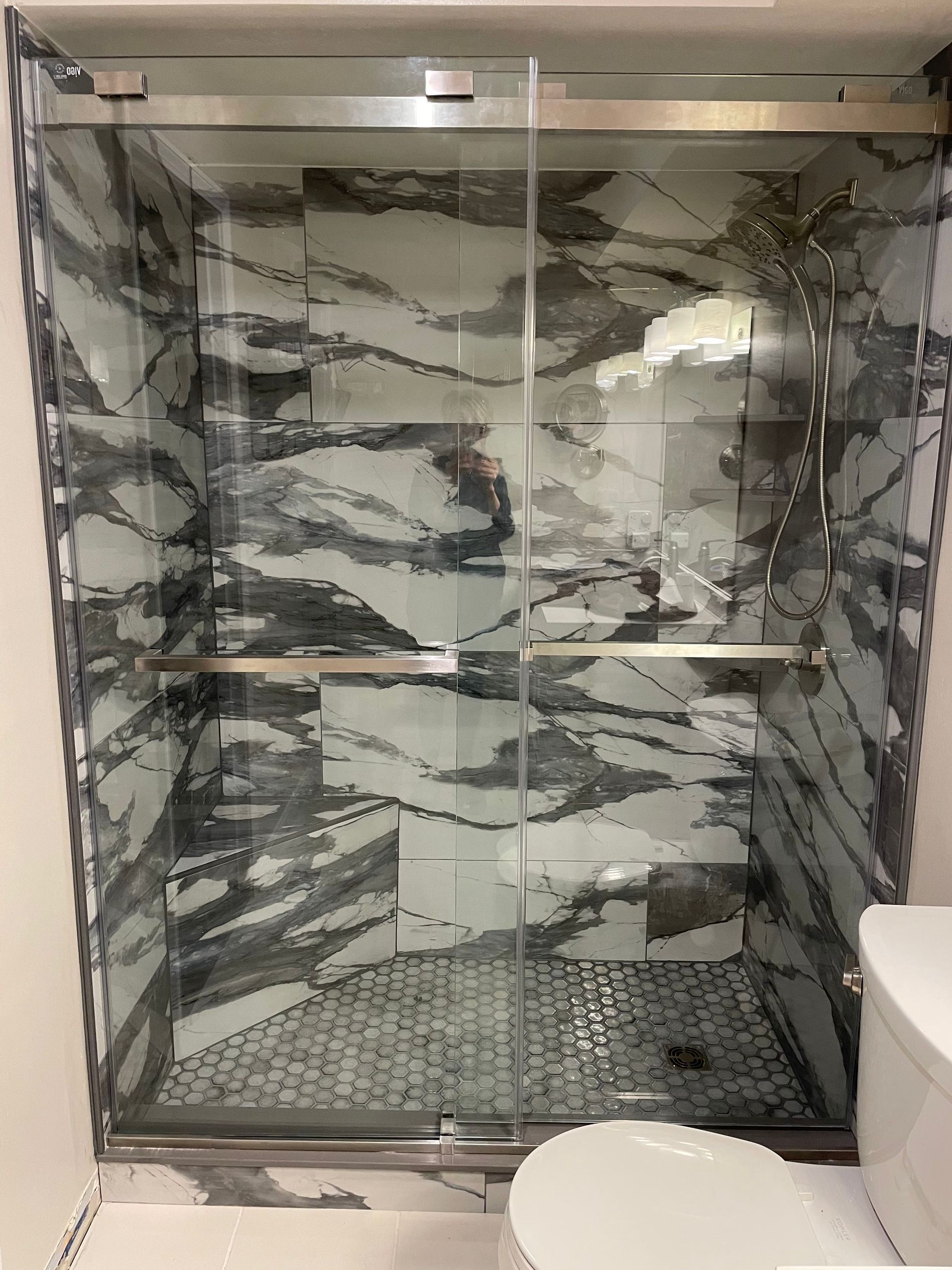 Glass-doored shower with marble-patterned walls and floor. Chrome fixtures. White toilet is to the right.