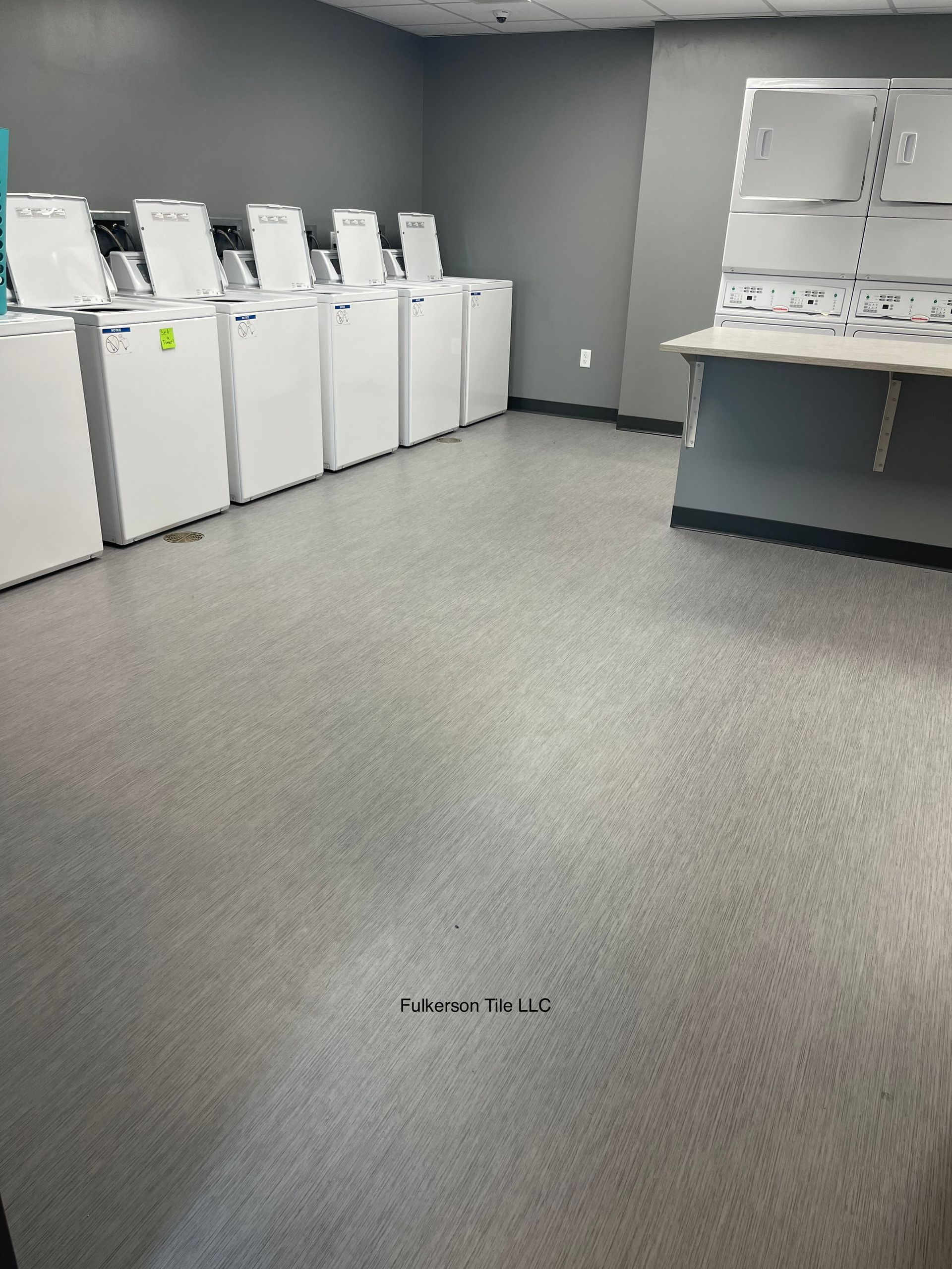 Row of white washing machines in a laundromat. Light gray floor and walls. A countertop on the right.