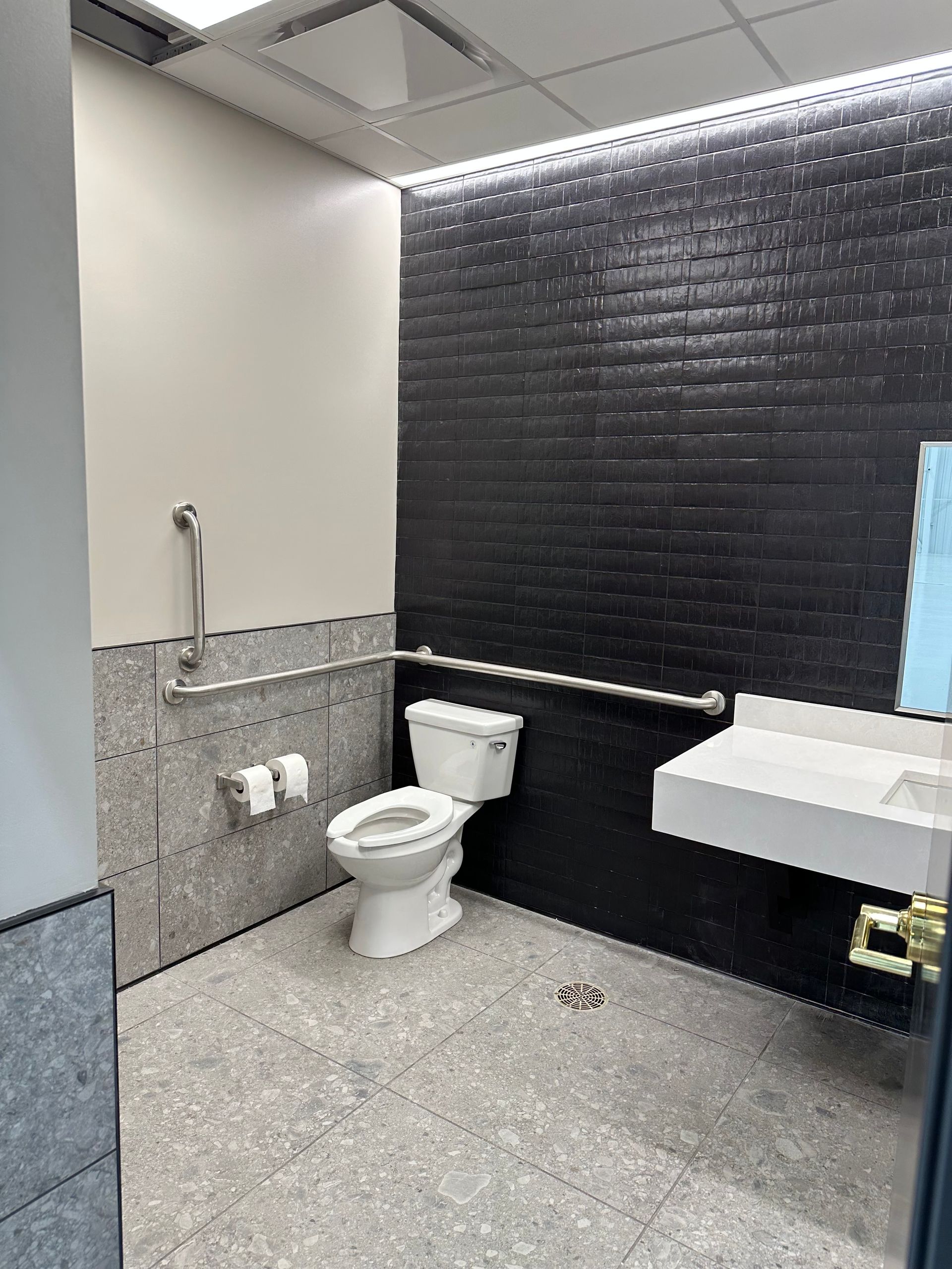 Accessible restroom with toilet, sink, and grab bars. Black brick accent wall, gray tile, white fixtures.
