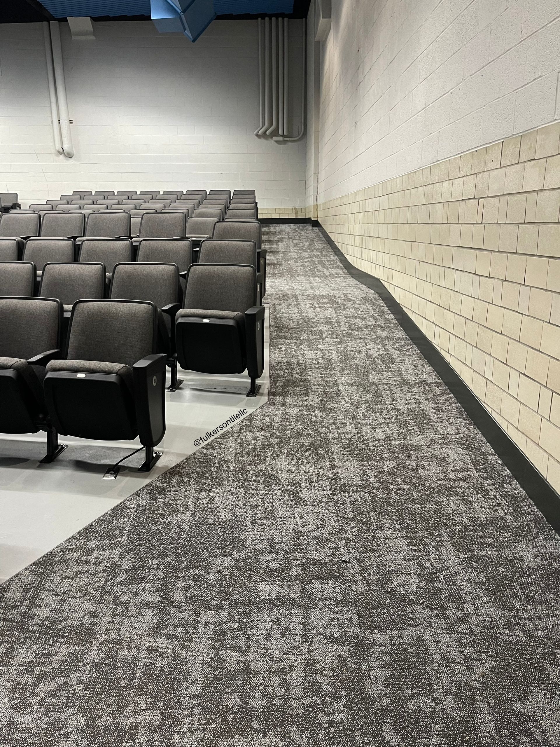 Auditorium seating with patterned carpeted aisle next to a white brick wall.