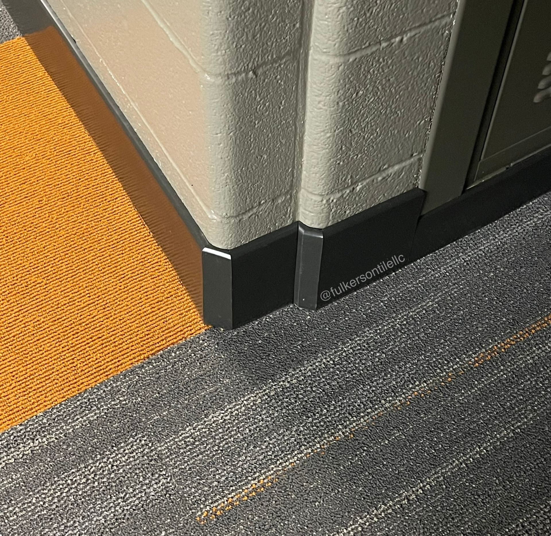 Corner of a beige brick wall and cabinet with dark baseboard, orange and gray carpet.