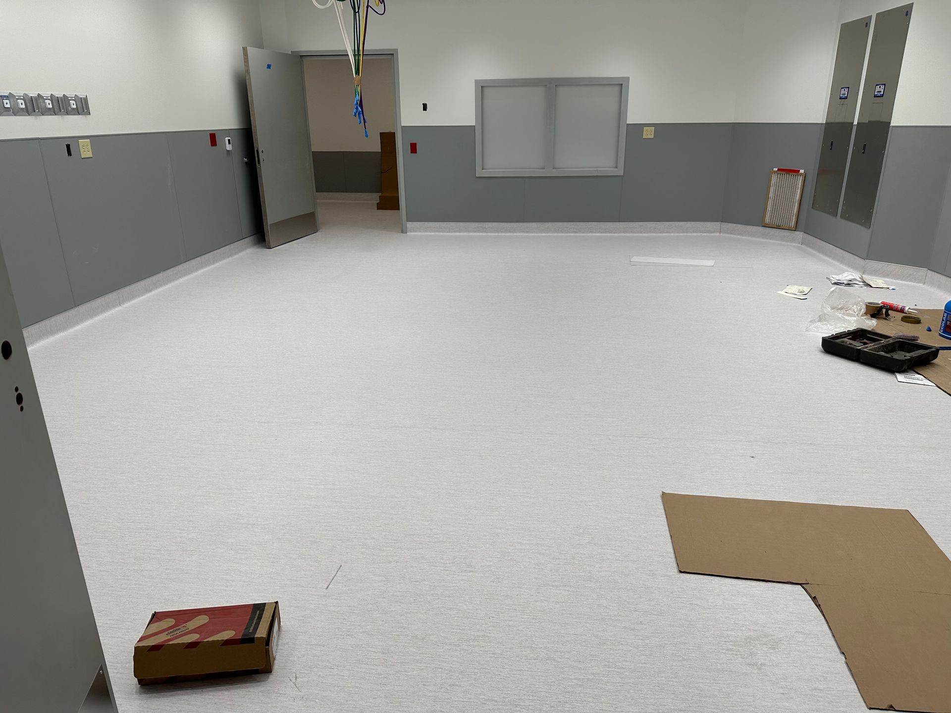 Empty room with light gray floor and walls; gray wainscoting; window and door visible. Construction materials on floor.