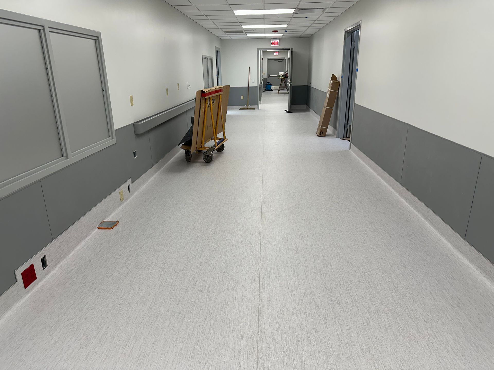 A long, empty hospital hallway with light gray walls, white upper walls, and a light gray speckled floor.