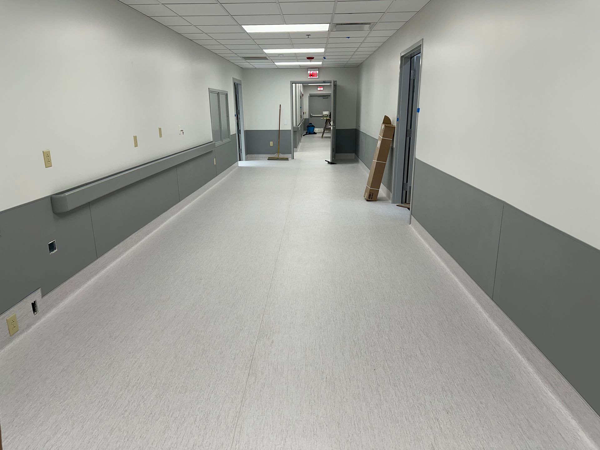 Hospital hallway with gray and white walls and flooring, handrail, and open doorways.