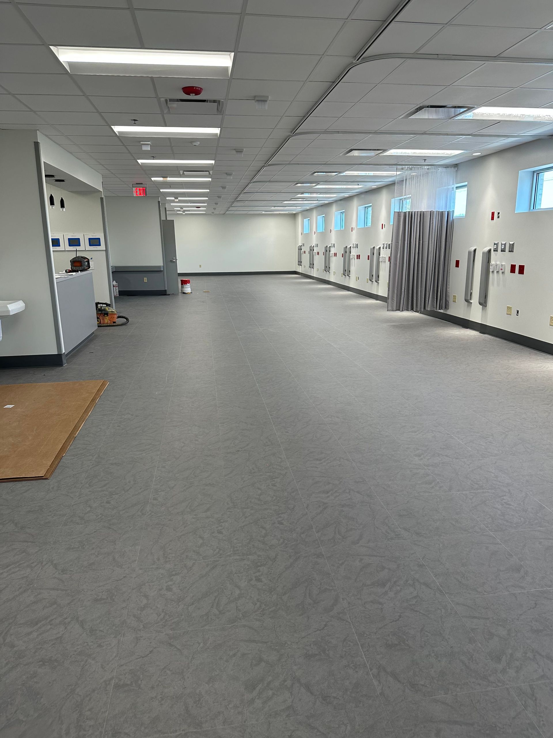 Long, empty room with grey flooring and white walls, medical fixtures along one wall.