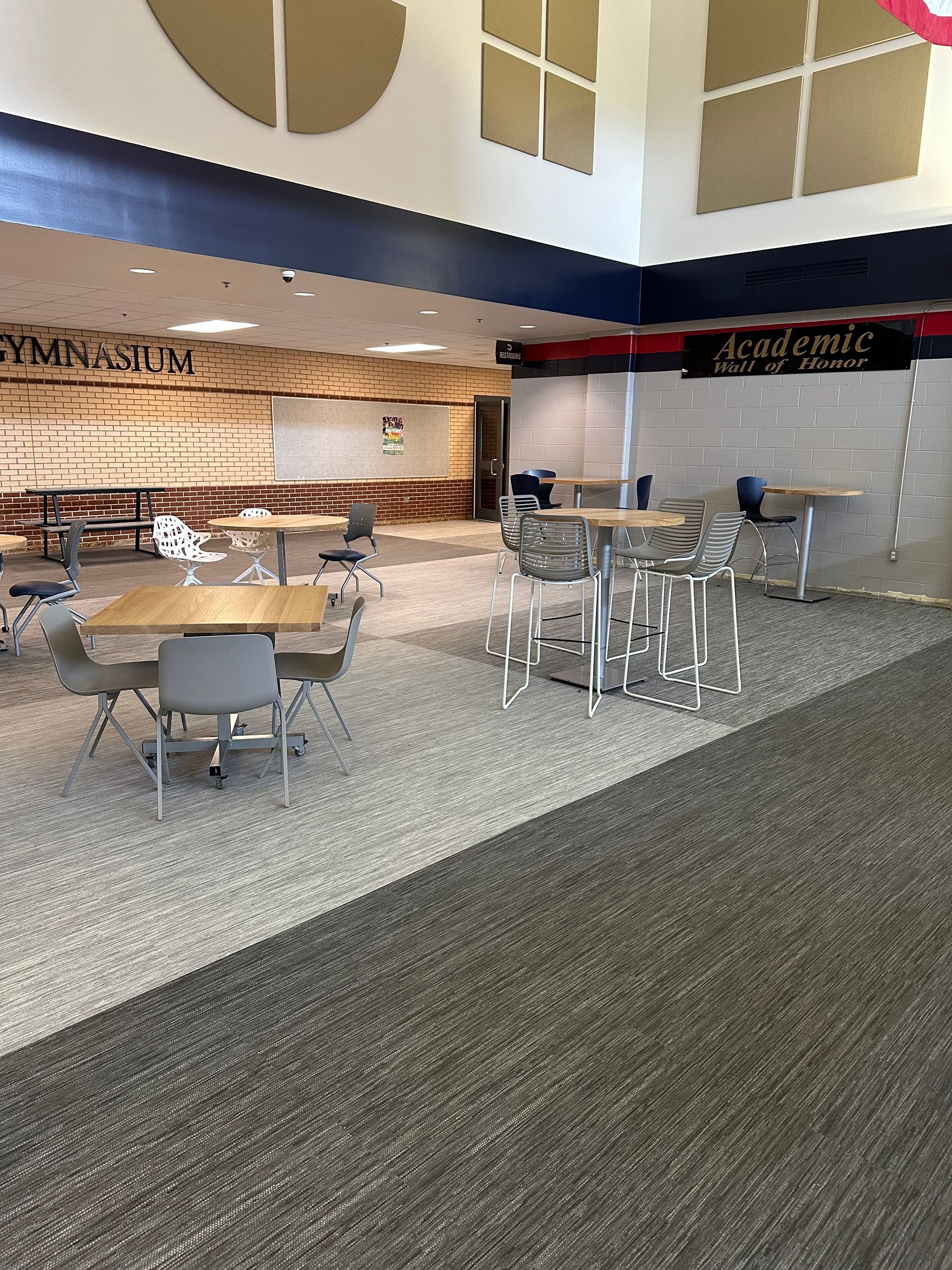 Open area with tables and chairs, possibly a school cafeteria. Gray and beige tones, natural light.