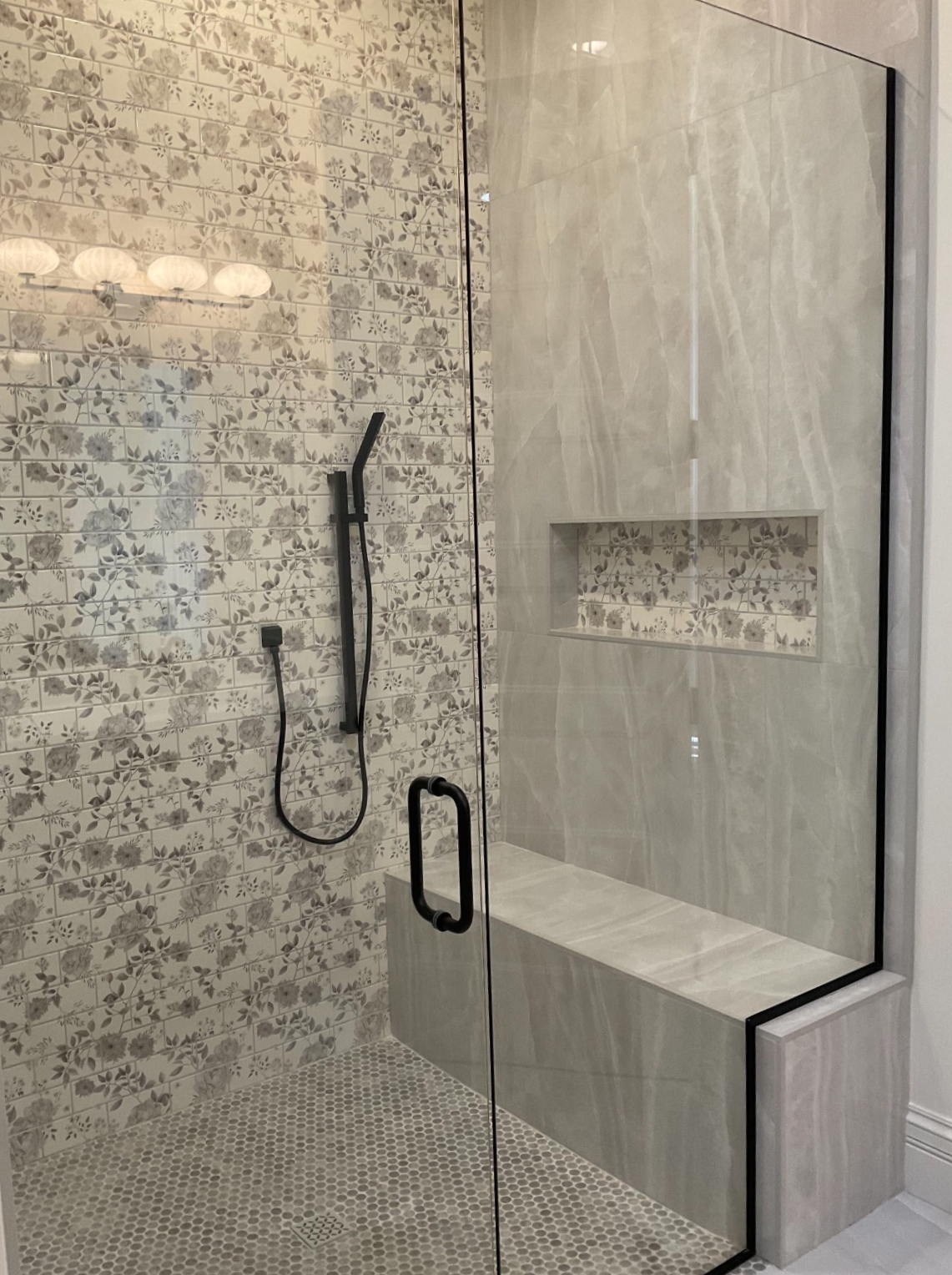 Shower with glass door, floral tile, built-in bench, dark fixtures, and a recessed shelf.