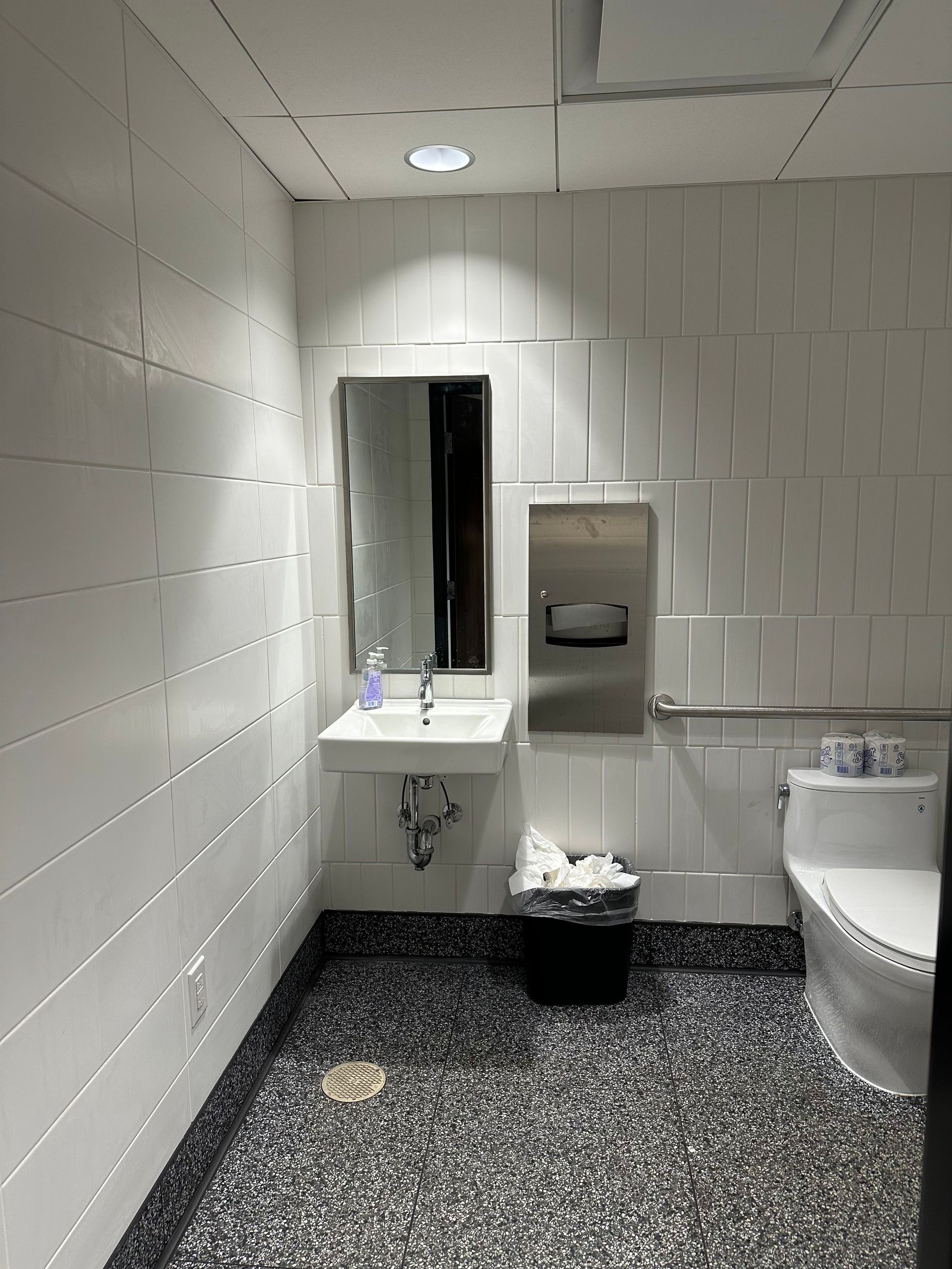 White-tiled restroom with a sink, mirror, toilet, and trash can; black speckled floor; stainless steel paper towel dispenser.
