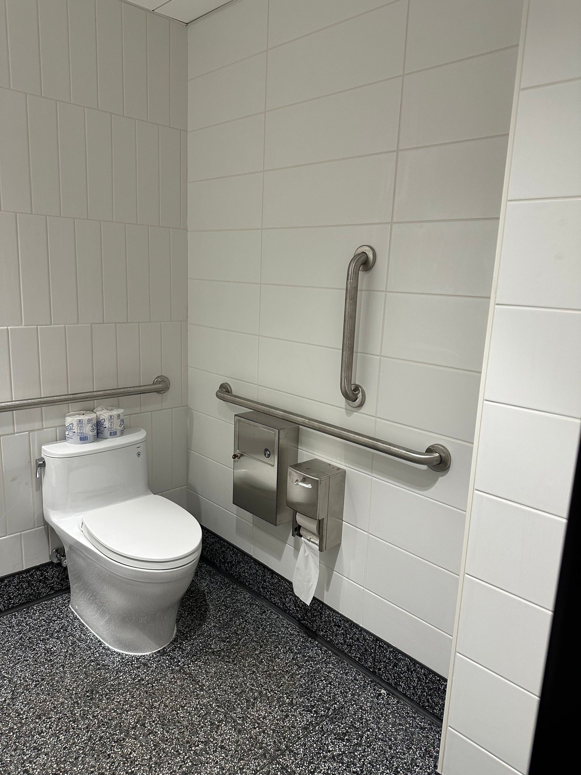 Accessible restroom with toilet, grab bars, and paper dispenser, white tiled walls, and speckled floor.