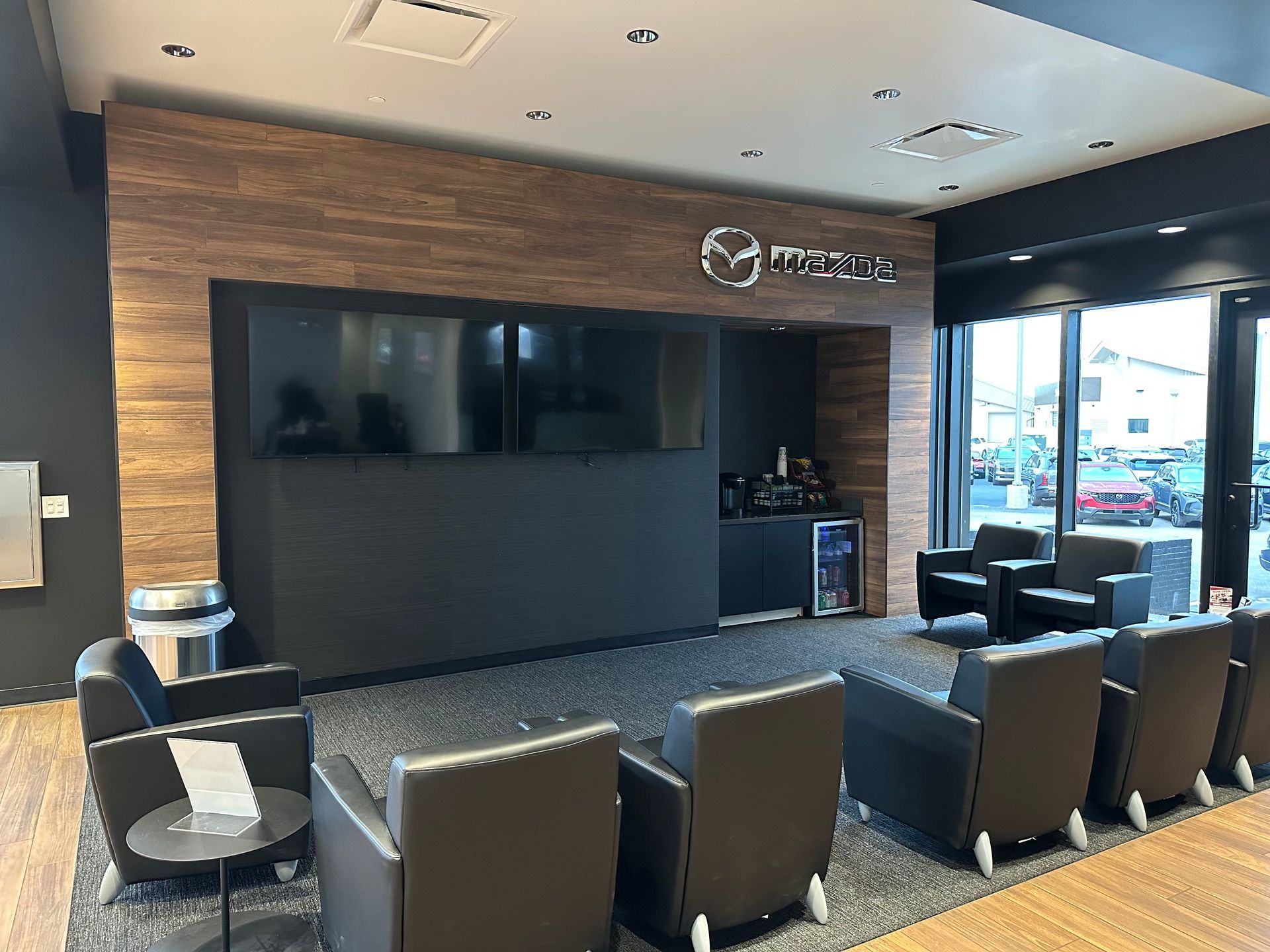 Mazda dealership waiting area with black chairs, TV screens, and a coffee station.