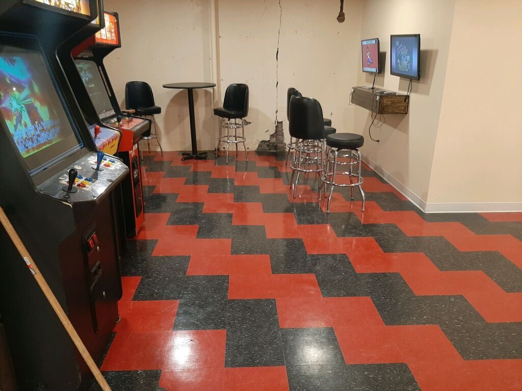 Arcade room with retro games, black and red zig-zag floor, stools, and monitors on the wall.