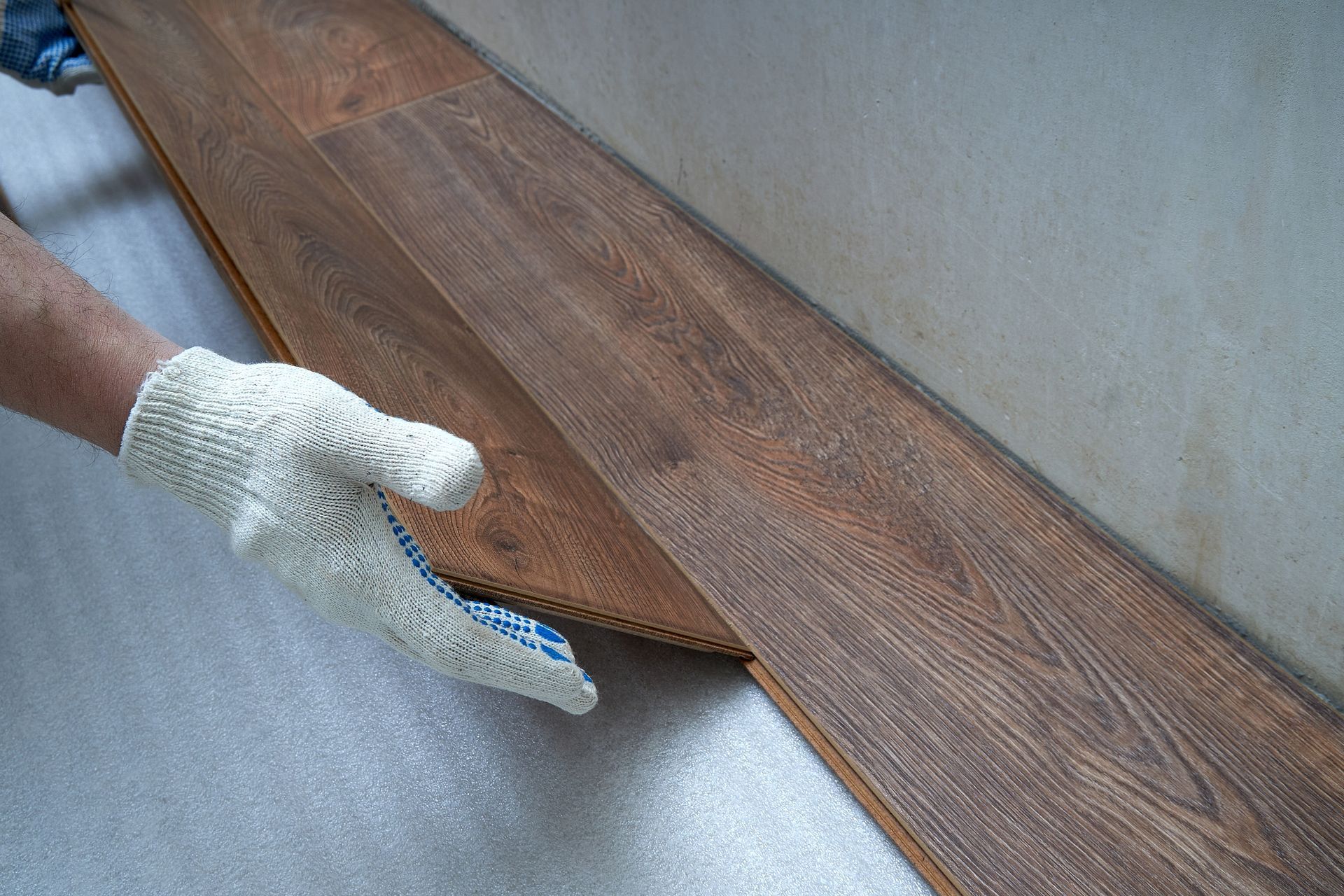 Person wearing a glove installing laminate flooring; brown planks against a gray surface and wall.
