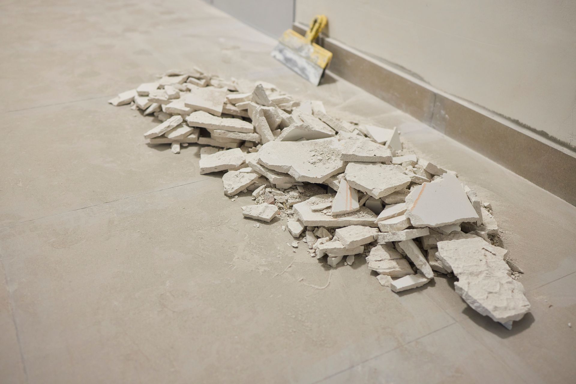 Pile of drywall debris on a light-colored floor, next to a wall. A putty knife rests on the wall.