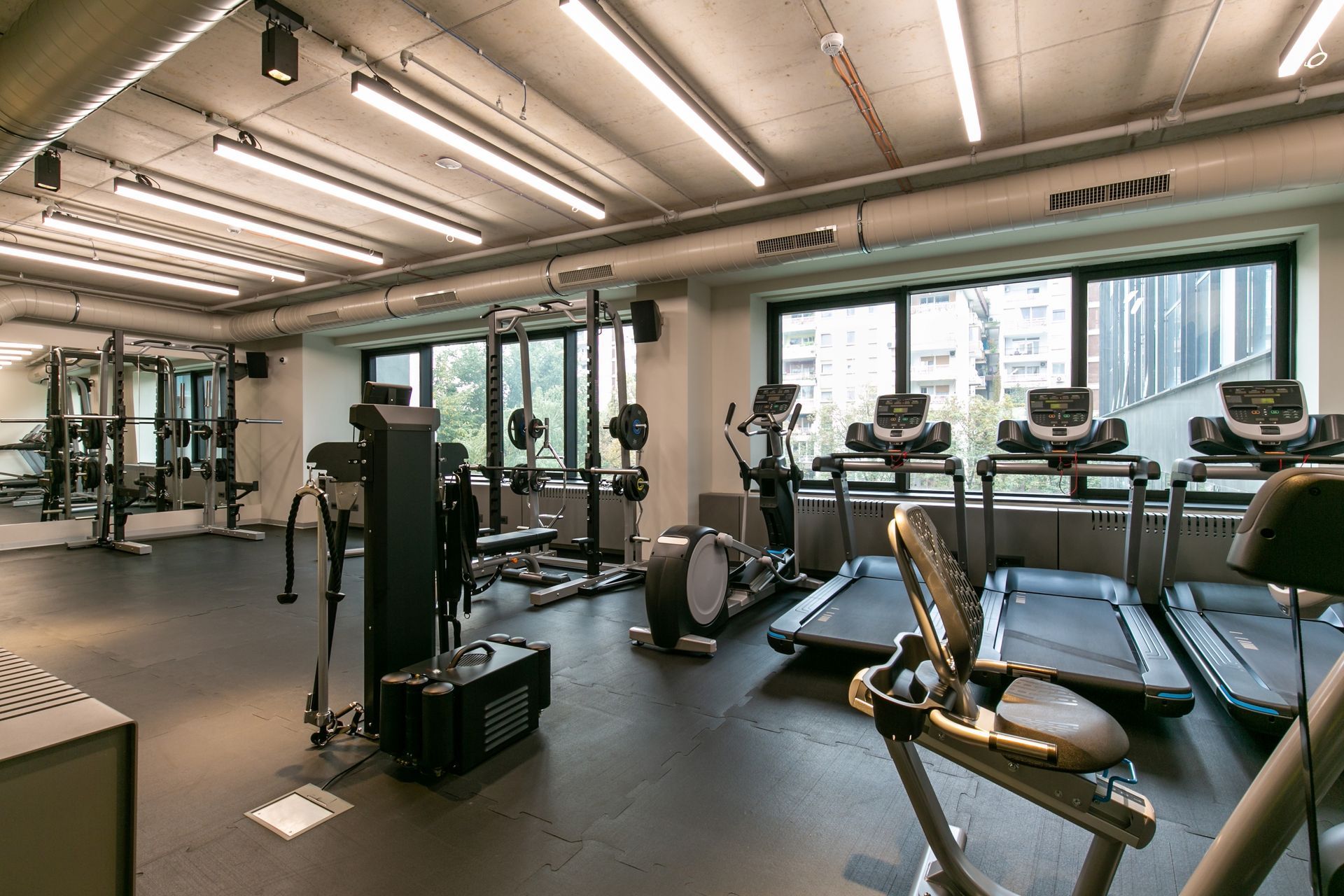 Gym with various exercise machines, including treadmills and weights, with large windows overlooking trees.