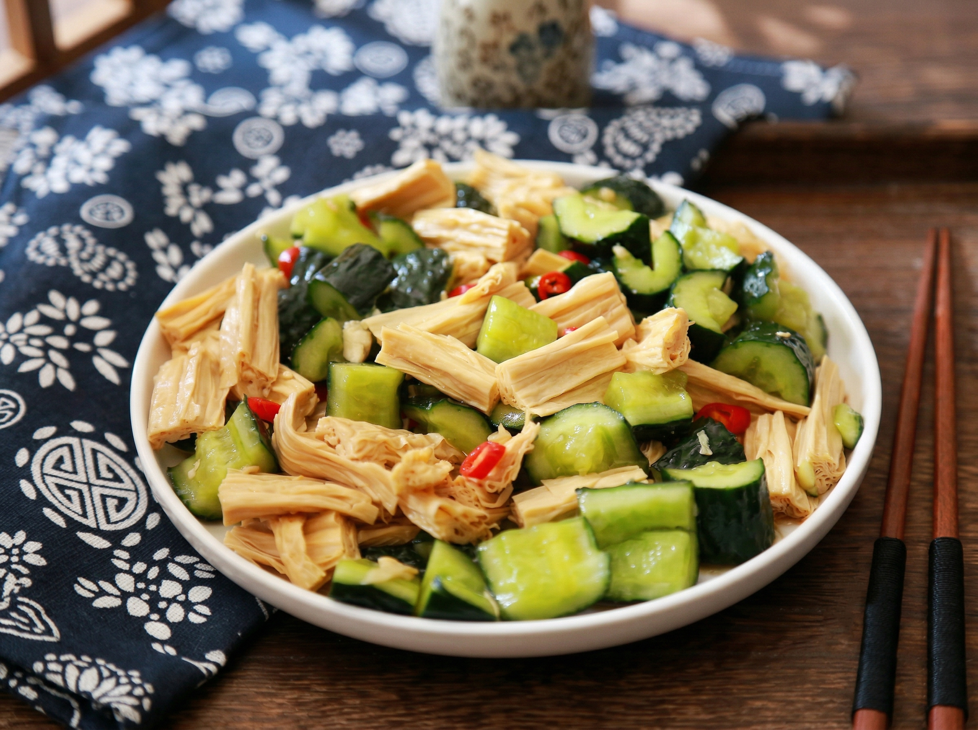 A white plate holds a mix of cucumber chunks and folded tofu skin strips on a dark blue patterned cloth and wooden table.
