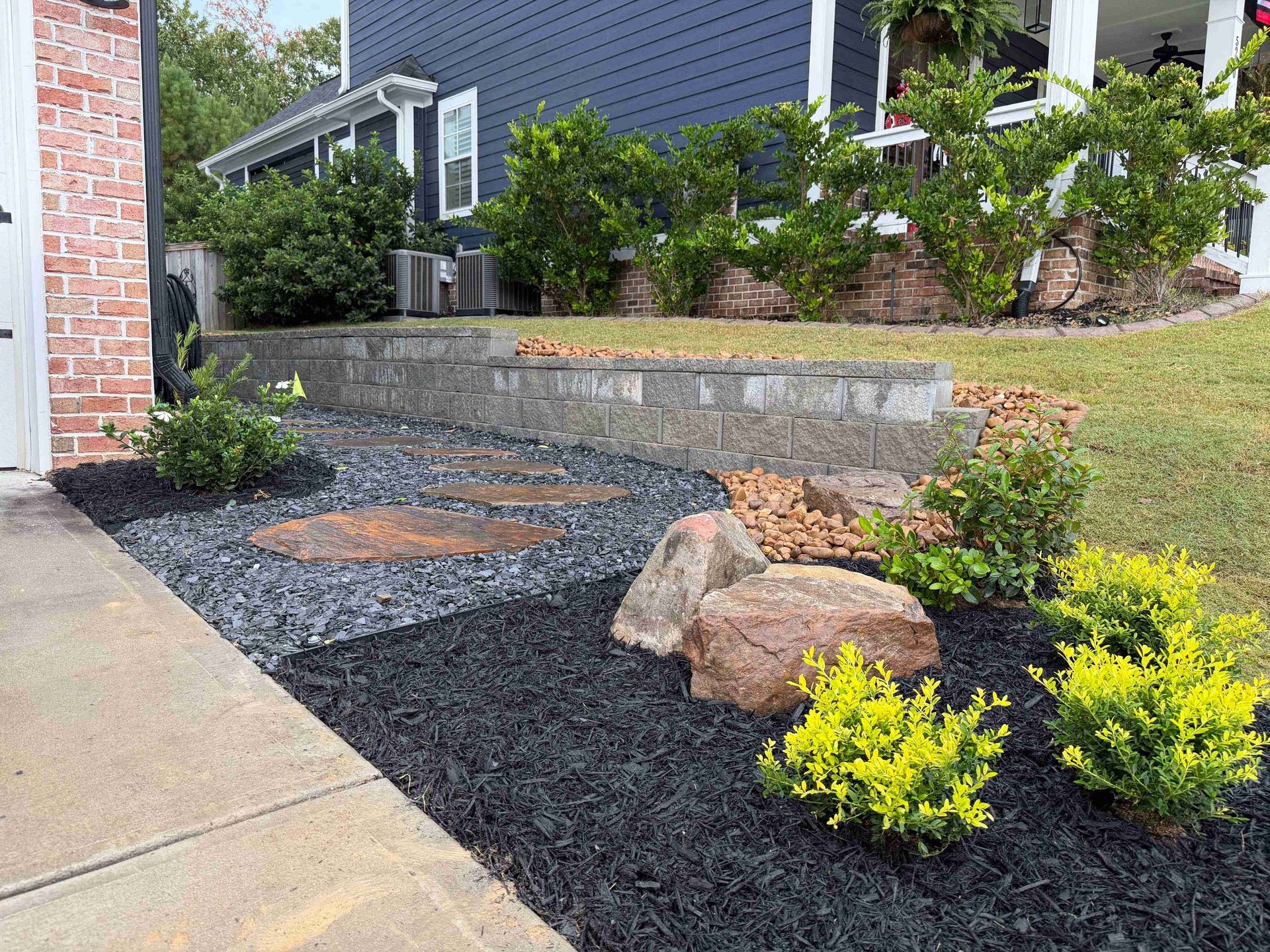 Landscaped front yard with dark mulch, stepping stones, rocks, and bushes in front of a blue house.