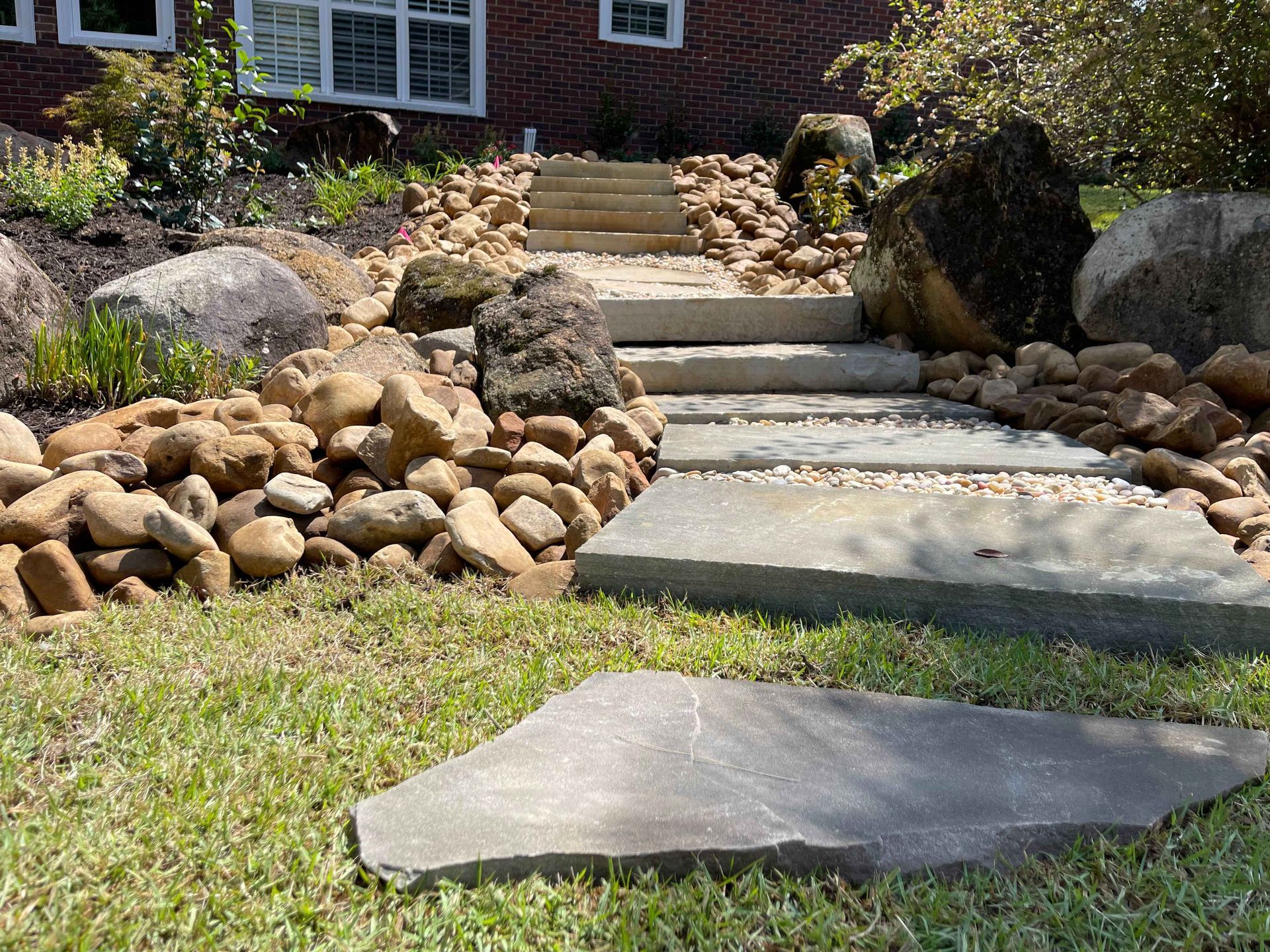 Stone pathway leading up steps, surrounded by rocks and landscaping, in front of a house.