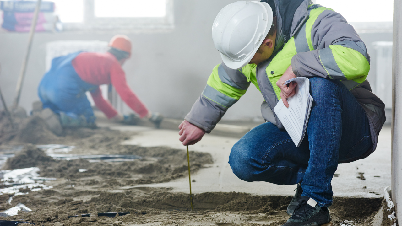 Construction worker in hard hat measures flooring, another works in background.