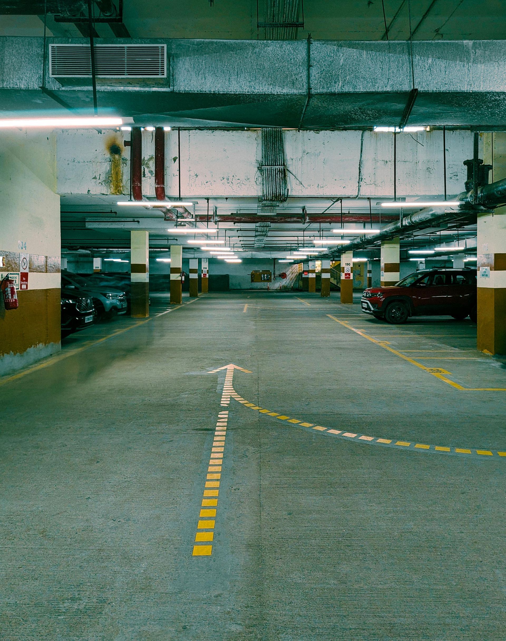 Parking garage interior with concrete floor, pillars, and parked cars. Yellow directional lines and overhead lighting.