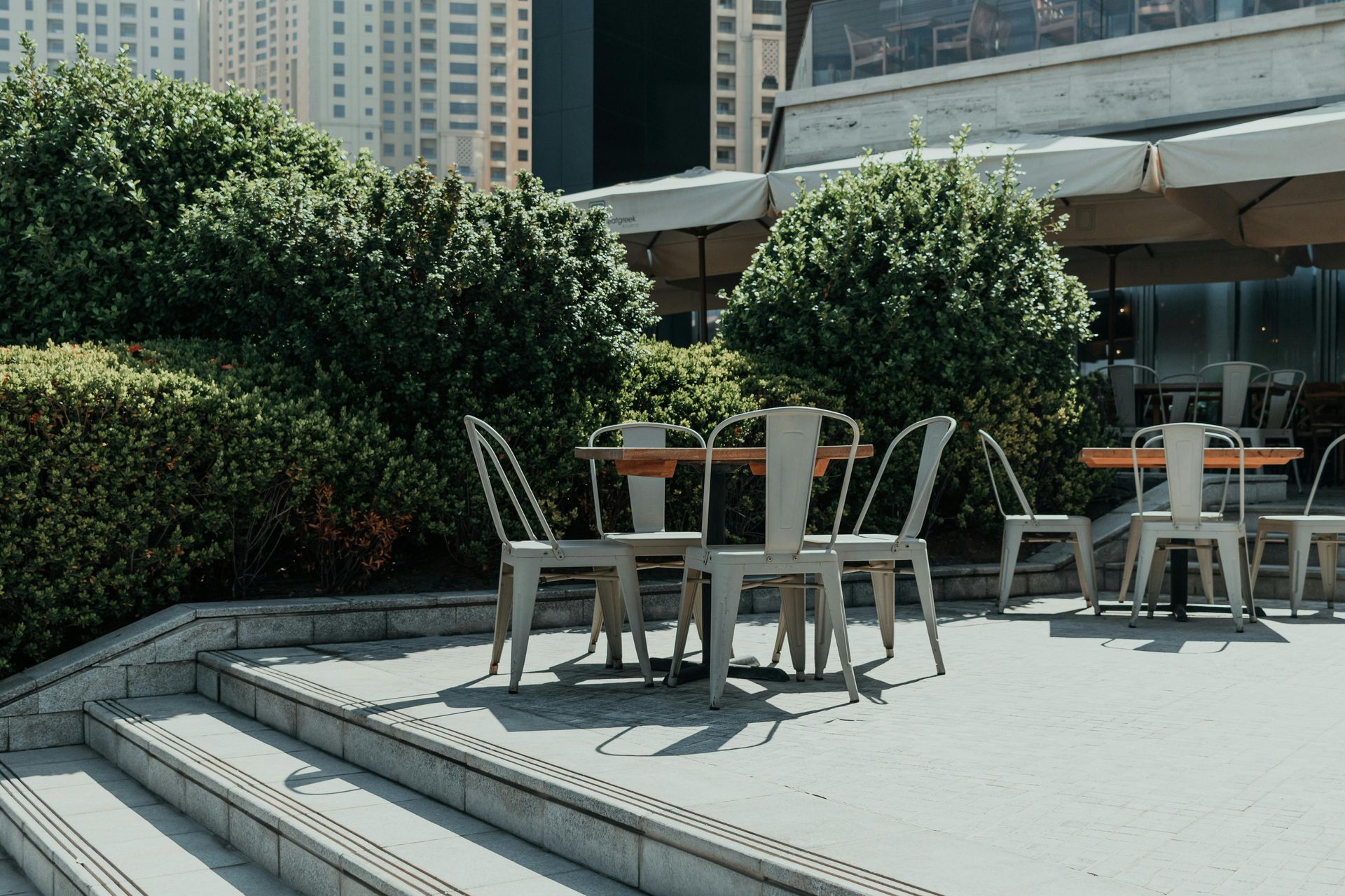 Outdoor cafe with tables and chairs on a patio, surrounded by green hedges and steps.