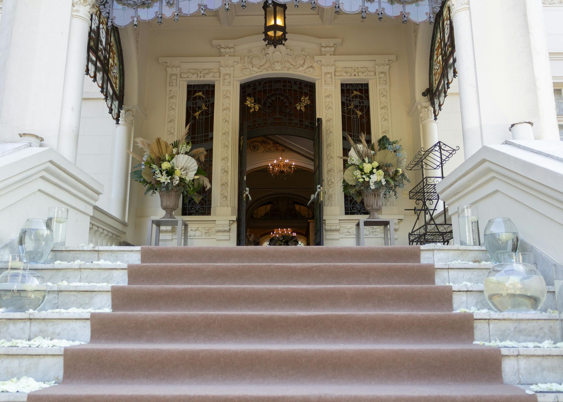 Exterior view of a grand mansion's entrance with steps leading to open double doors; floral arrangements flank the entrance.