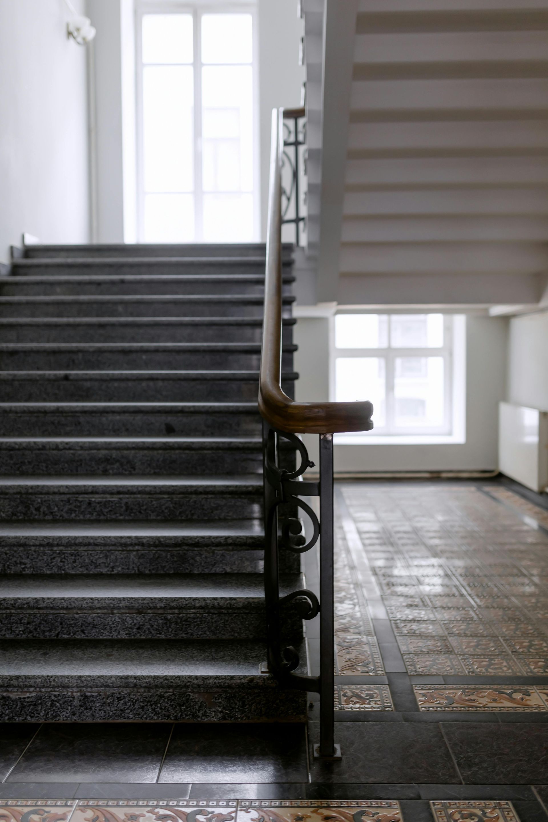 Staircase with gray steps, wooden handrail, and patterned tile floor in a building. Two windows visible.