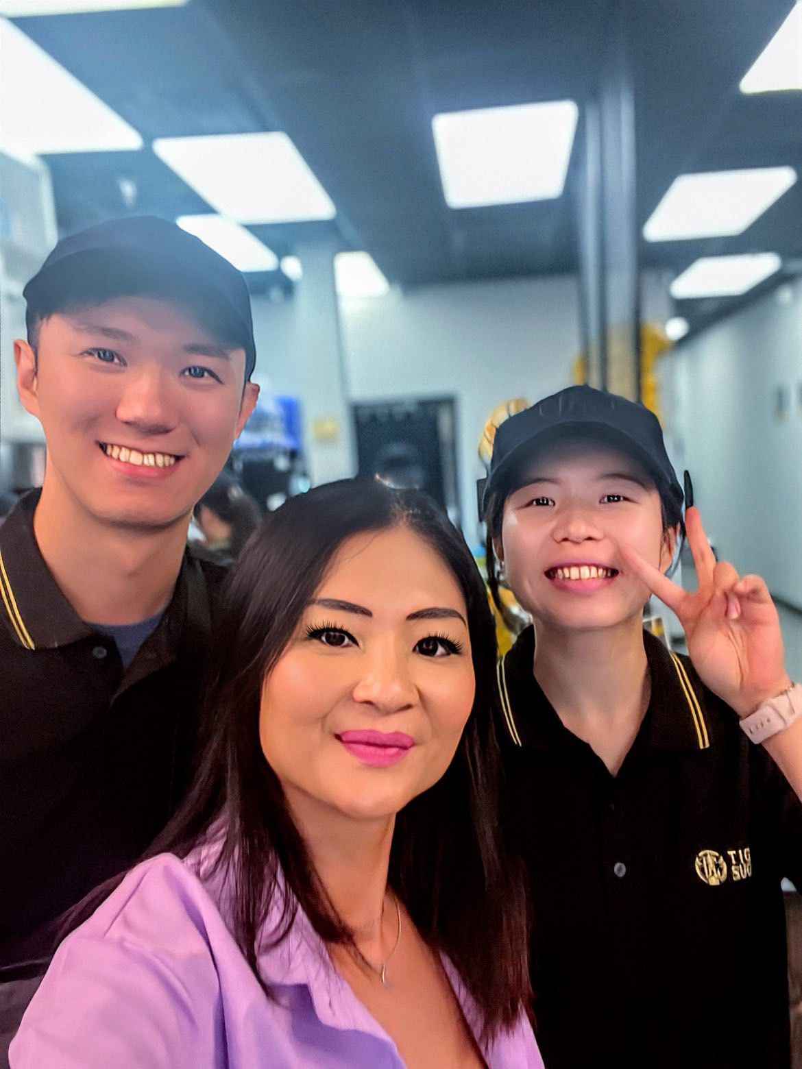 Three people smiling indoors, two in black uniforms, one in a purple shirt. Fluorescent lights above.