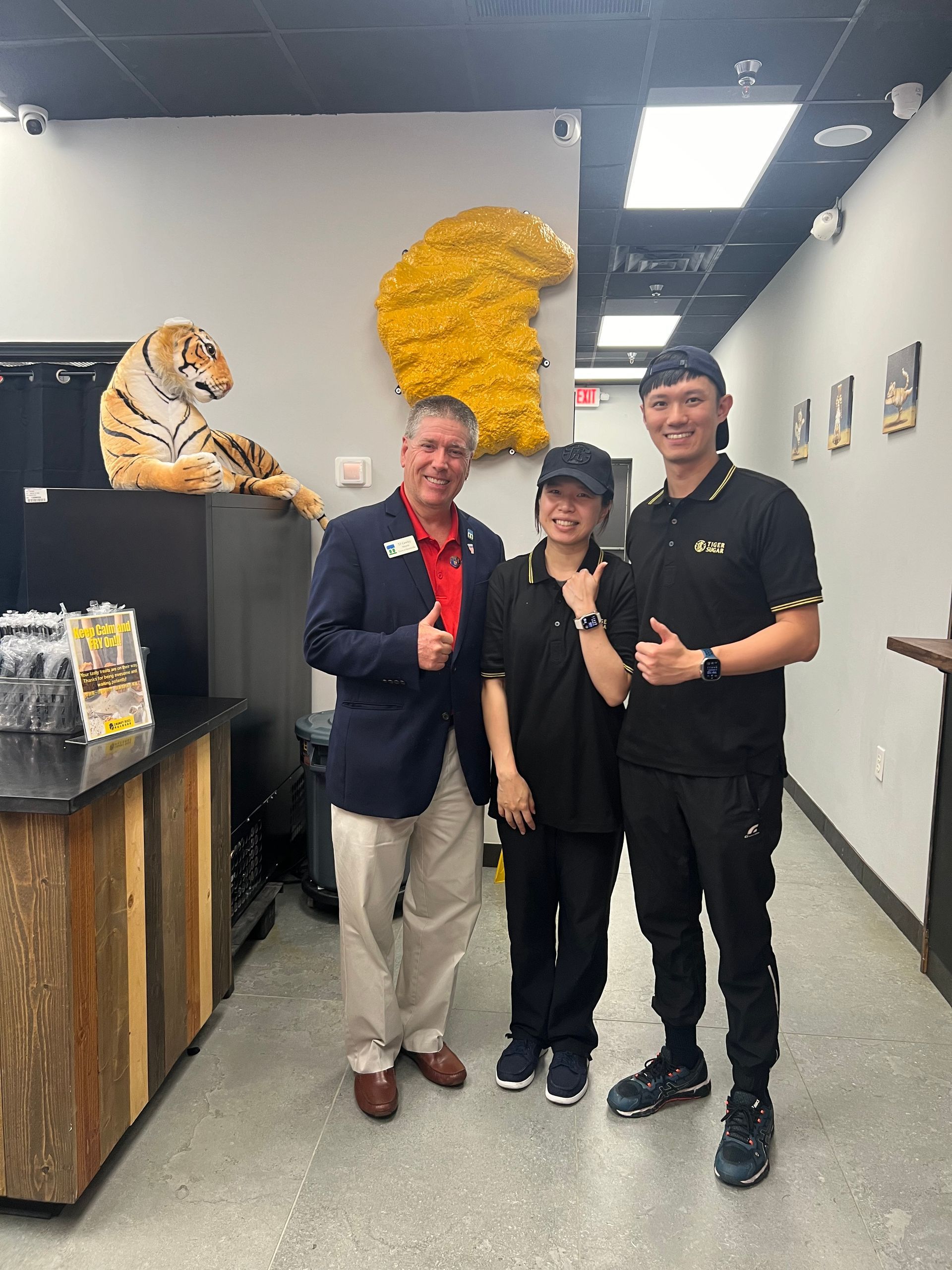 Man in blazer with two employees at a restaurant counter, giving thumbs up. Tiger statue and wall art visible.