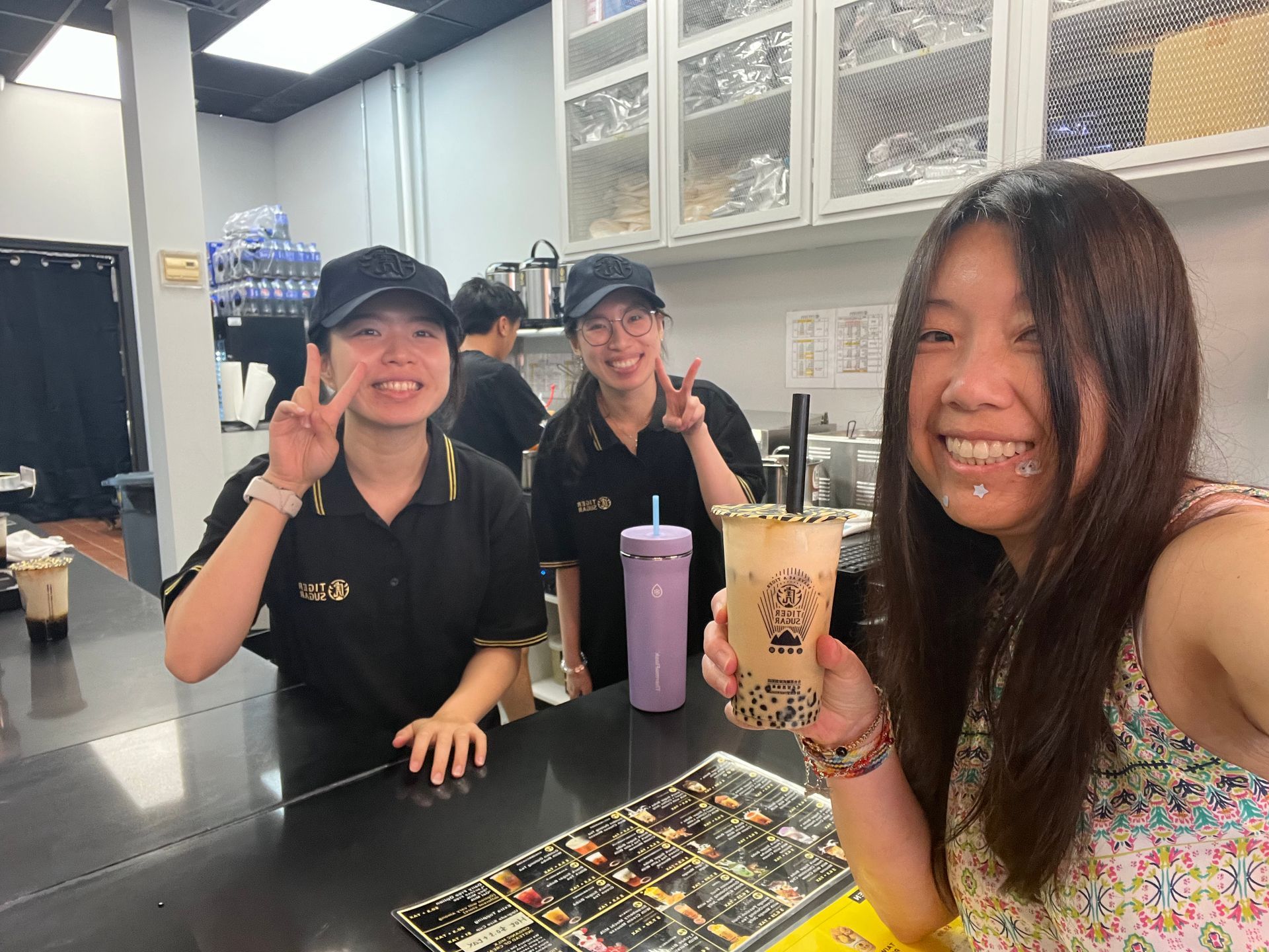 Woman smiles, holds boba tea, poses with two employees in black uniforms, behind a counter.