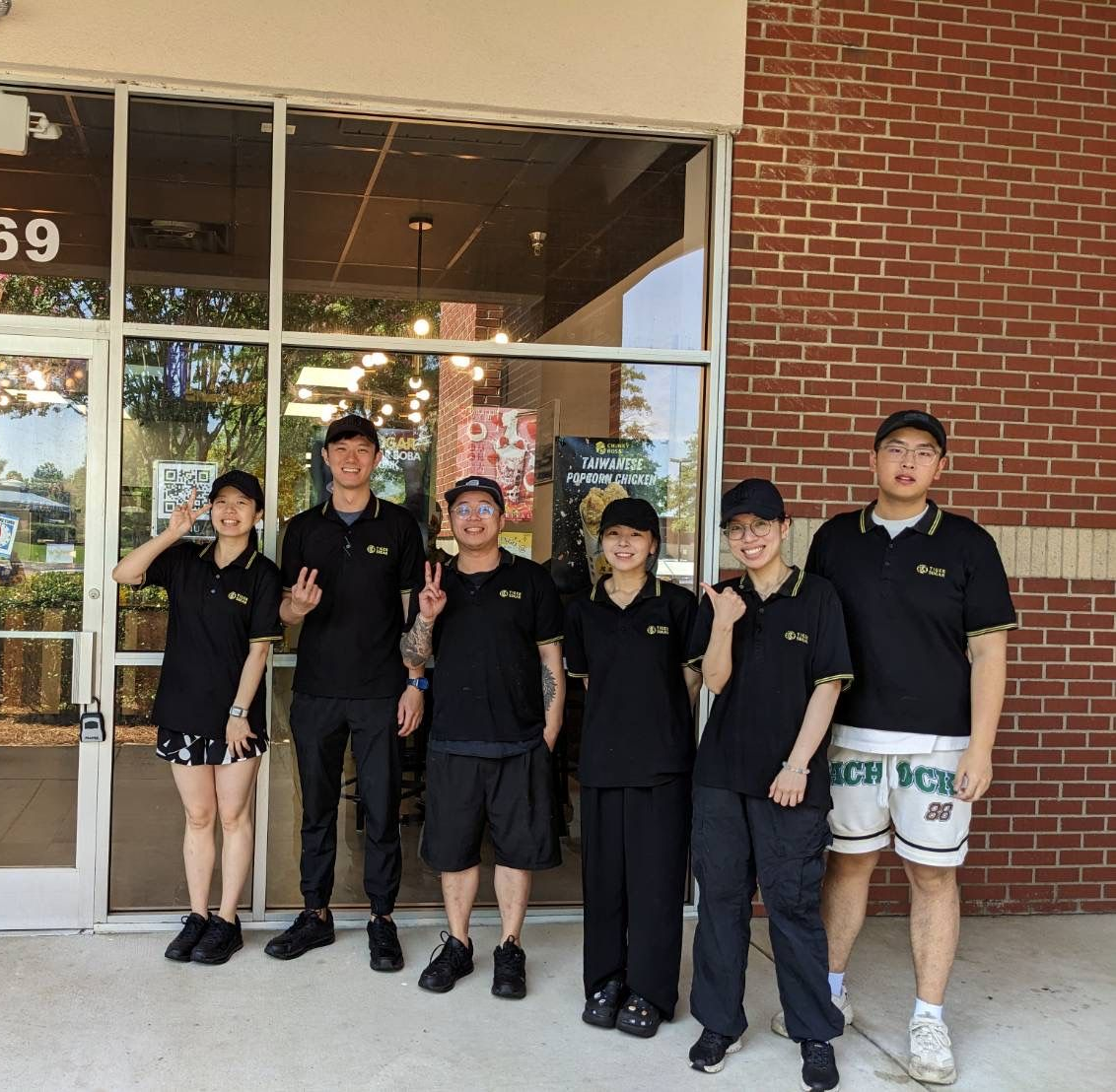 Group of seven people in black uniforms outside a brick building. Some are smiling, giving peace signs.