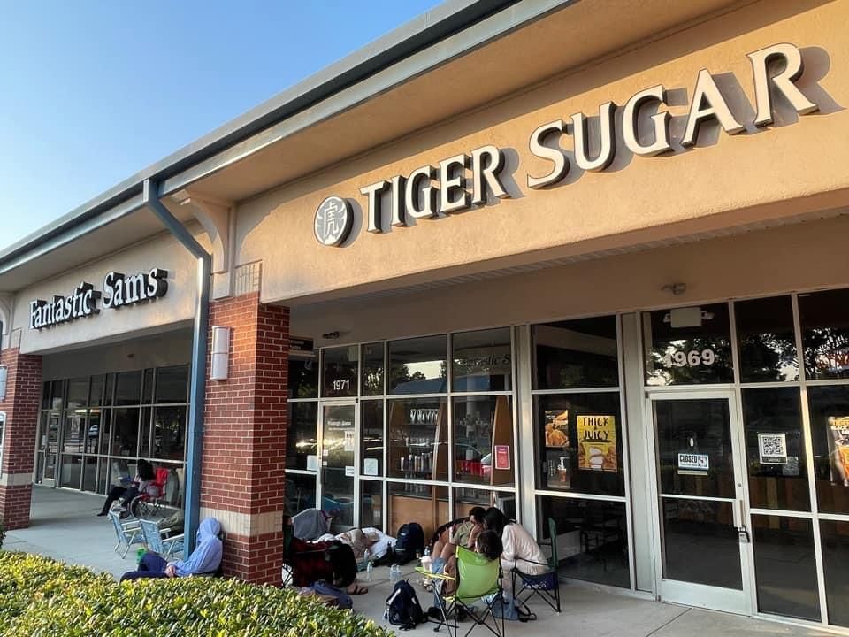 People sit outside of Tiger Sugar, waiting in line. The store has large windows and a sign with its name above the entrance.