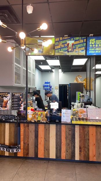 A bubble tea shop counter with two workers behind it, facing the customer, in a brightly lit space. The counter has a wooden facade.