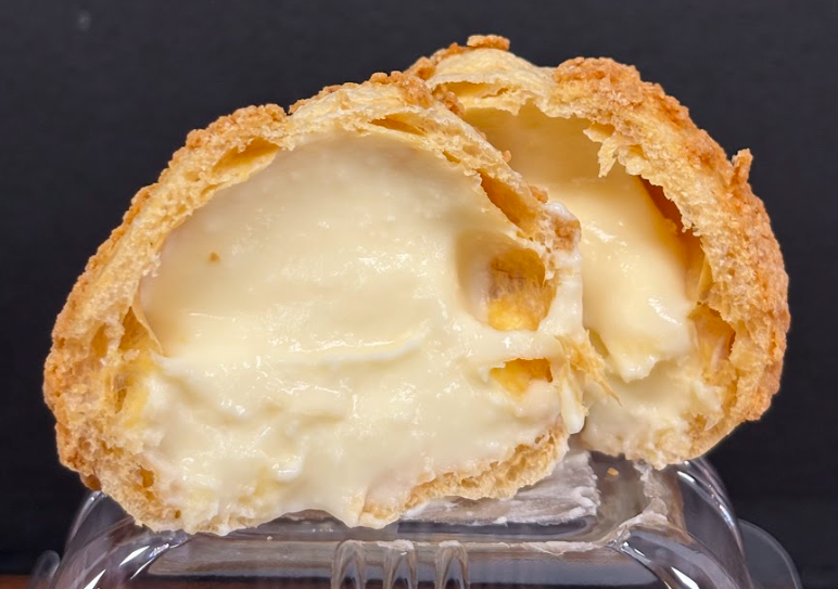 Cream puff cut in half, revealing a creamy custard filling. The golden pastry is resting on a plastic container.