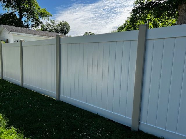 A white vinyl fence with brown posts is sitting in the grass in front of a house.