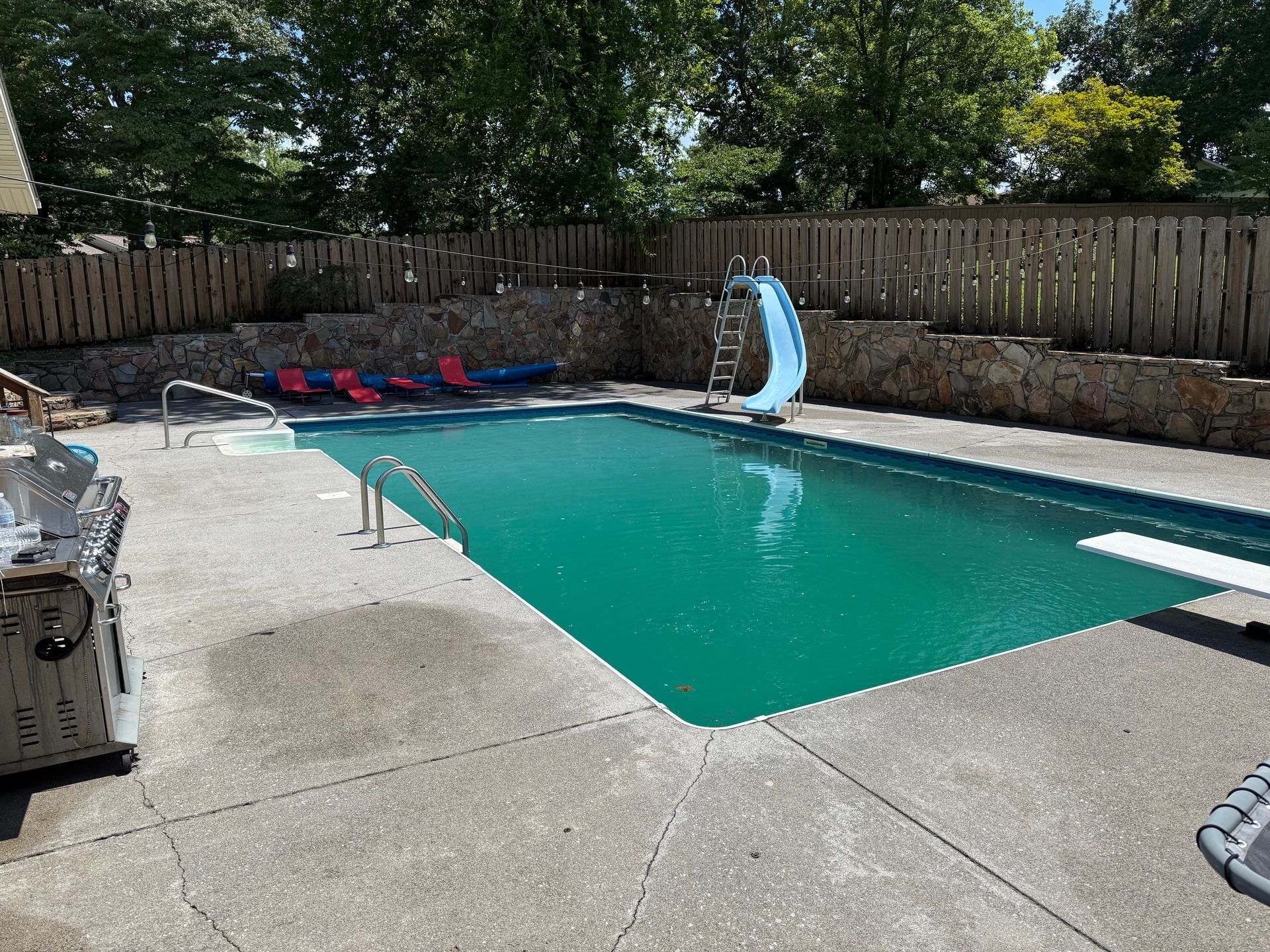 Pool with a slide and diving board surrounded by a stone wall and wooden fence.