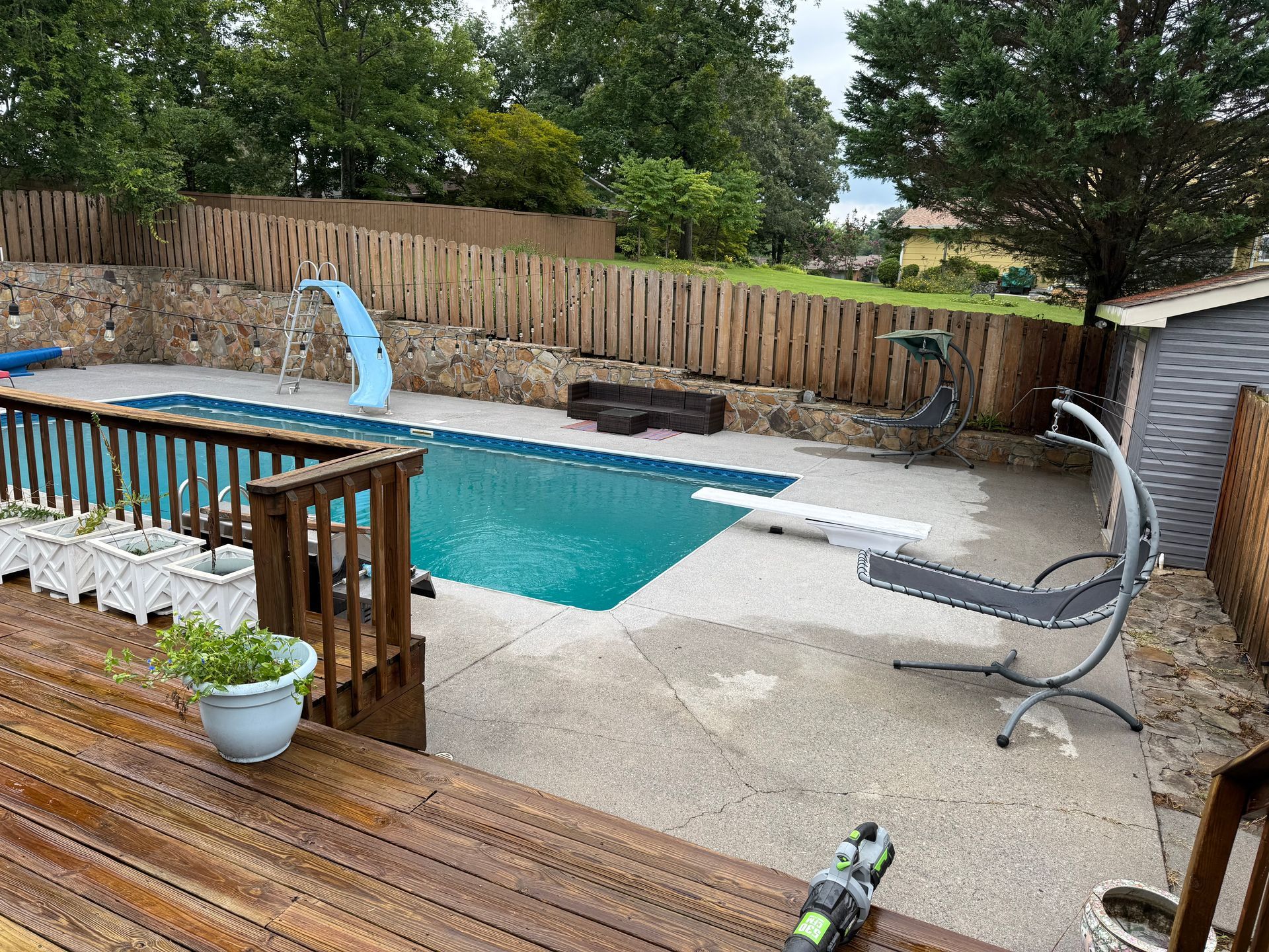 Backyard pool with a diving board, slide, wooden deck, and fence.