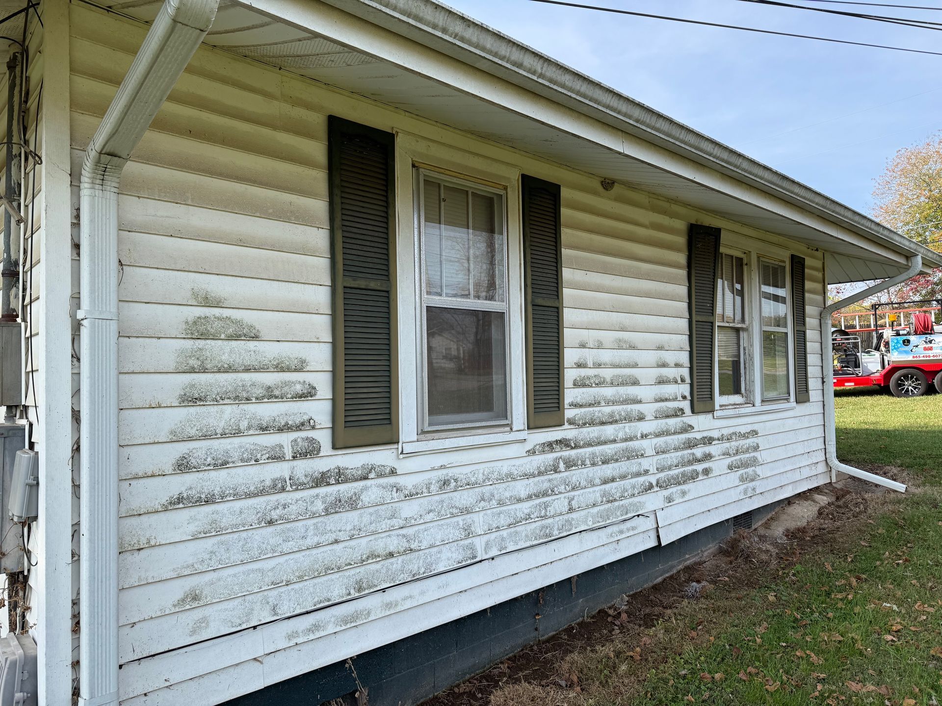 White house siding with dark shutters, covered in mold. Green grass and blue sky are visible.